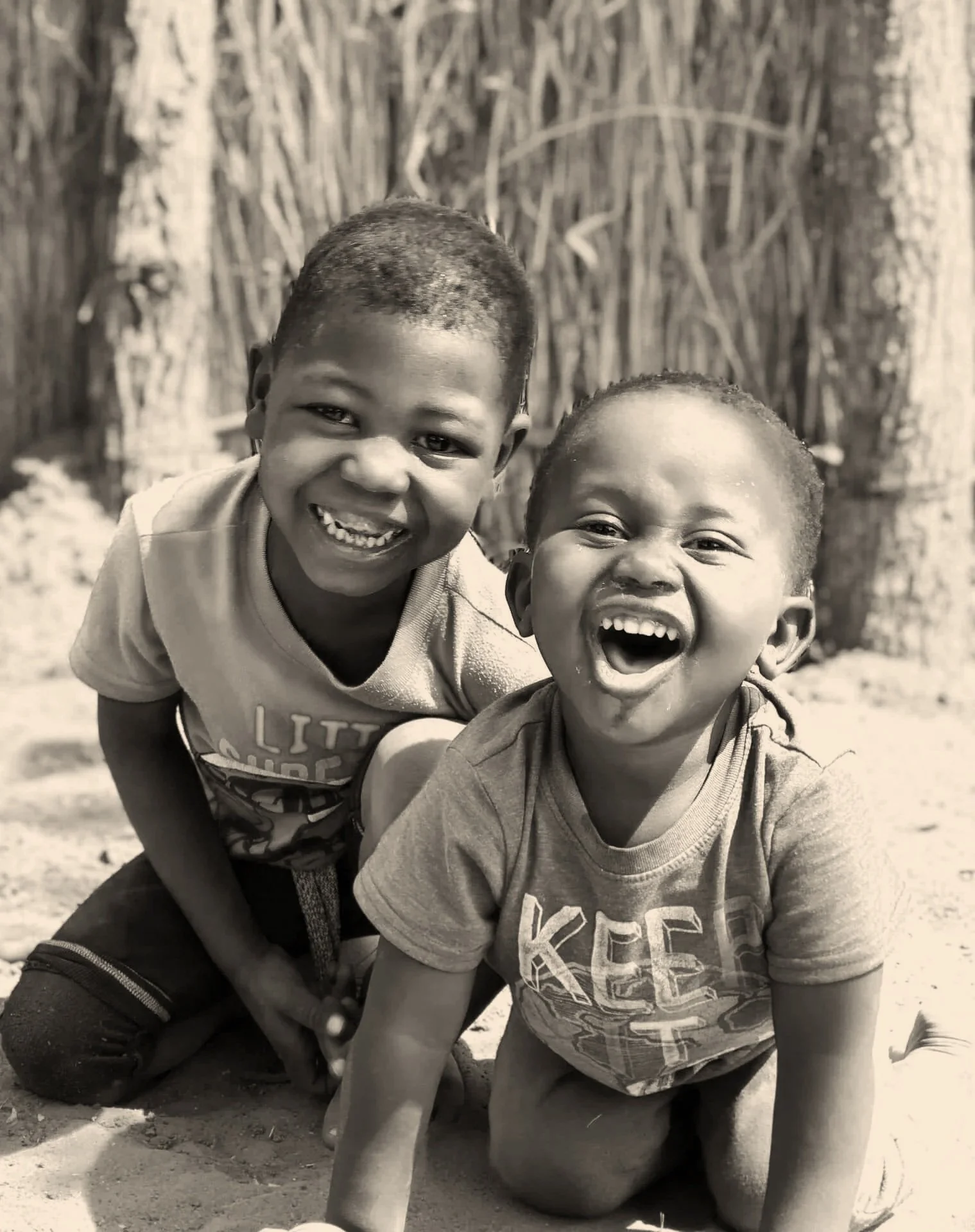 Two boys laughing and playing outdoors on the ground with trees in the background.