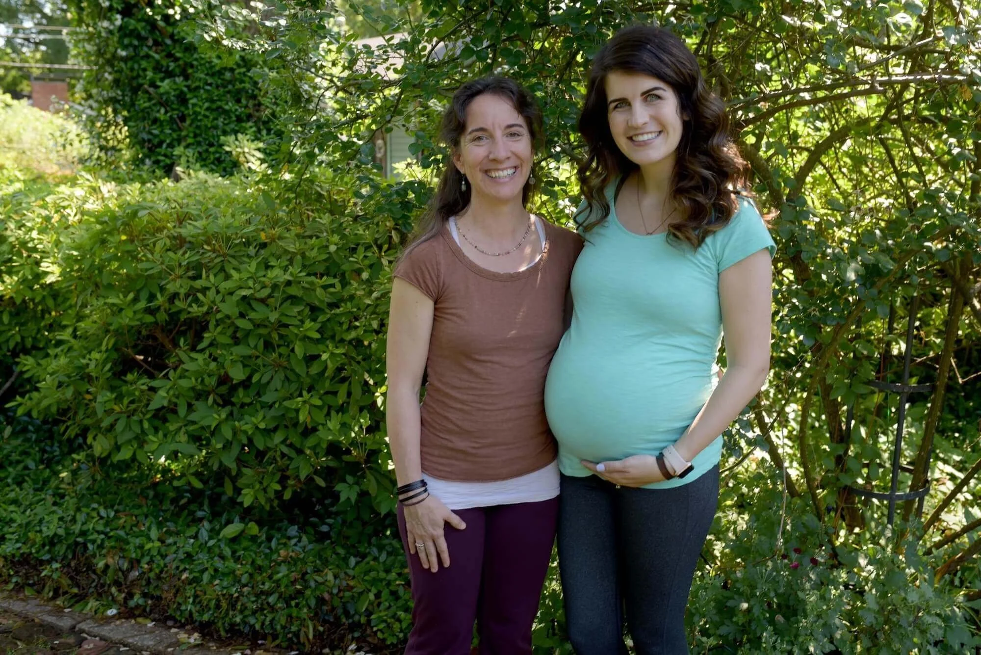 A pregnant woman with long, curly brown hair wearing a light blue shirt and black pants standing outdoors next to a woman with long brown hair, wearing a brown shirt and purple pants, both smiling. They are in front of greenery and sunlight.