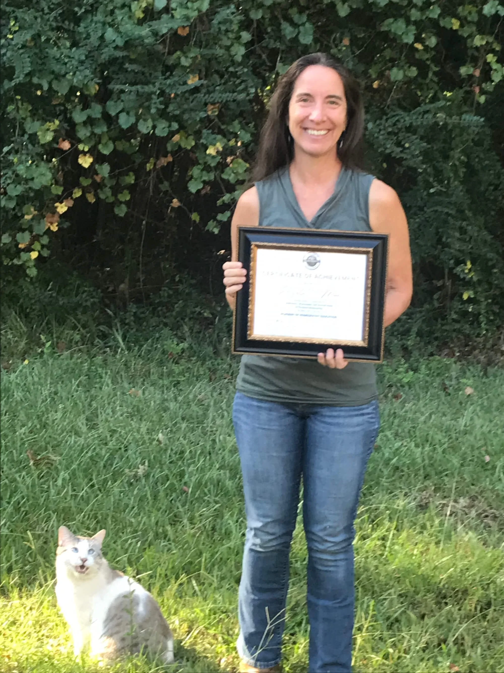 A woman holding a framed certificate standing on grass with a cat sitting on the grass nearby, in front of dense green foliage.