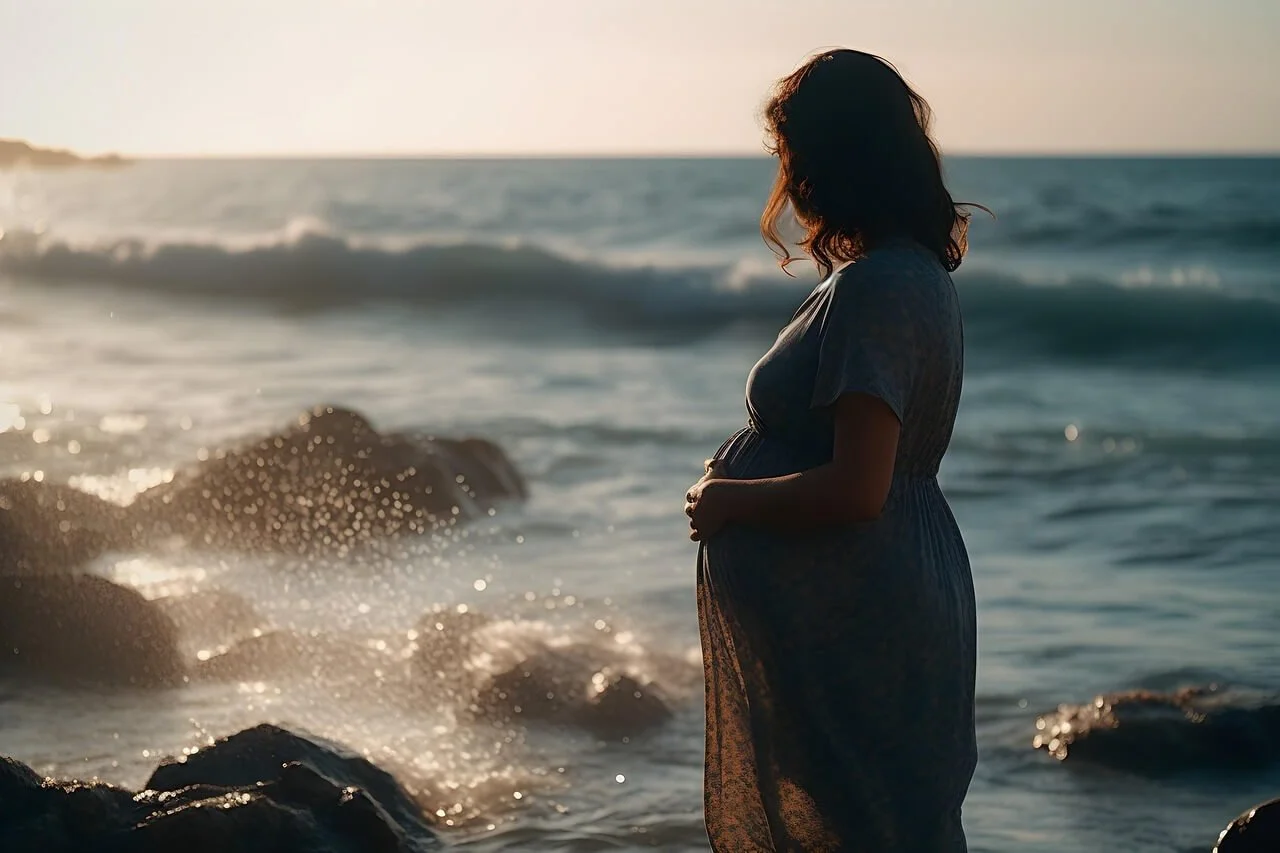 A pregnant woman standing on the beach during sunset, holding her belly and looking at the ocean with waves crashing on rocks in the background.