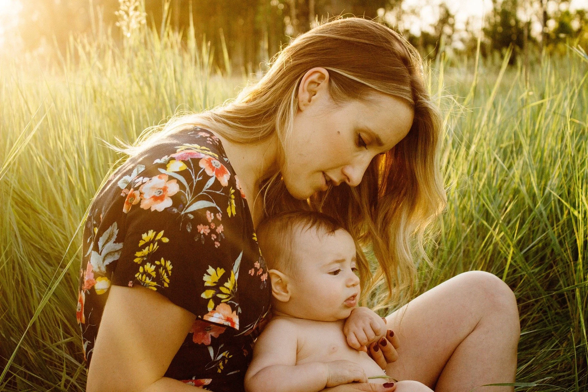 A woman with long, wavy blonde hair and a floral dress sitting with a baby in a grassy field during sunset.