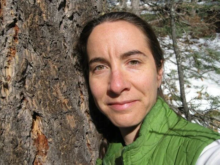 Woman with dark hair and green jacket standing next to a tree in a forested area.