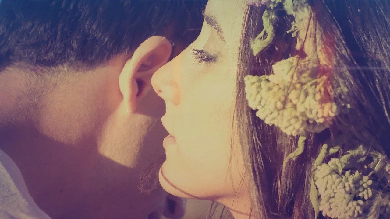 A close-up of a young couple with their faces close together, eyes closed, with the woman wearing flowers in her hair.