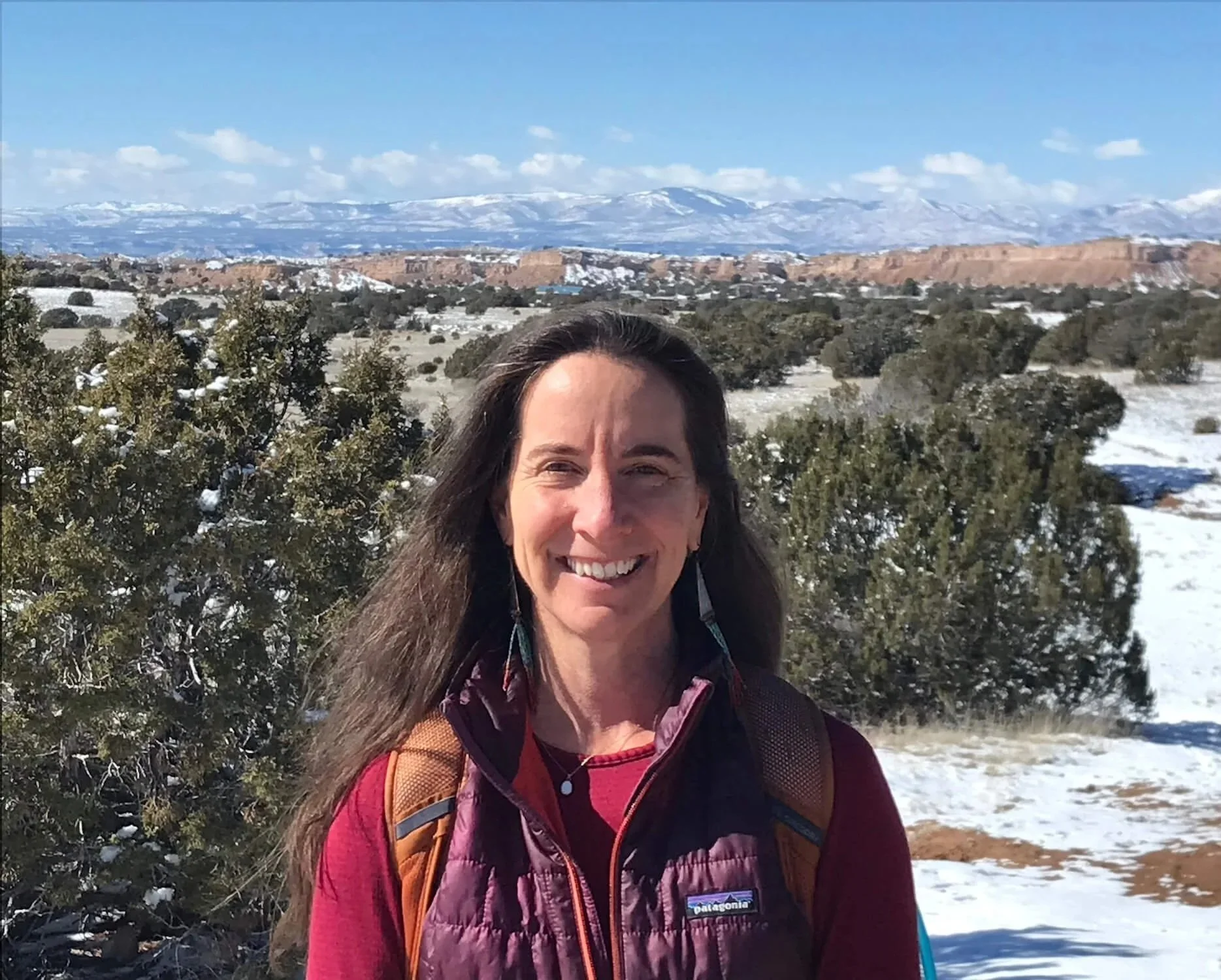 A woman smiling outdoors in snowy terrain with green bushes, mountains and blue sky in the background.