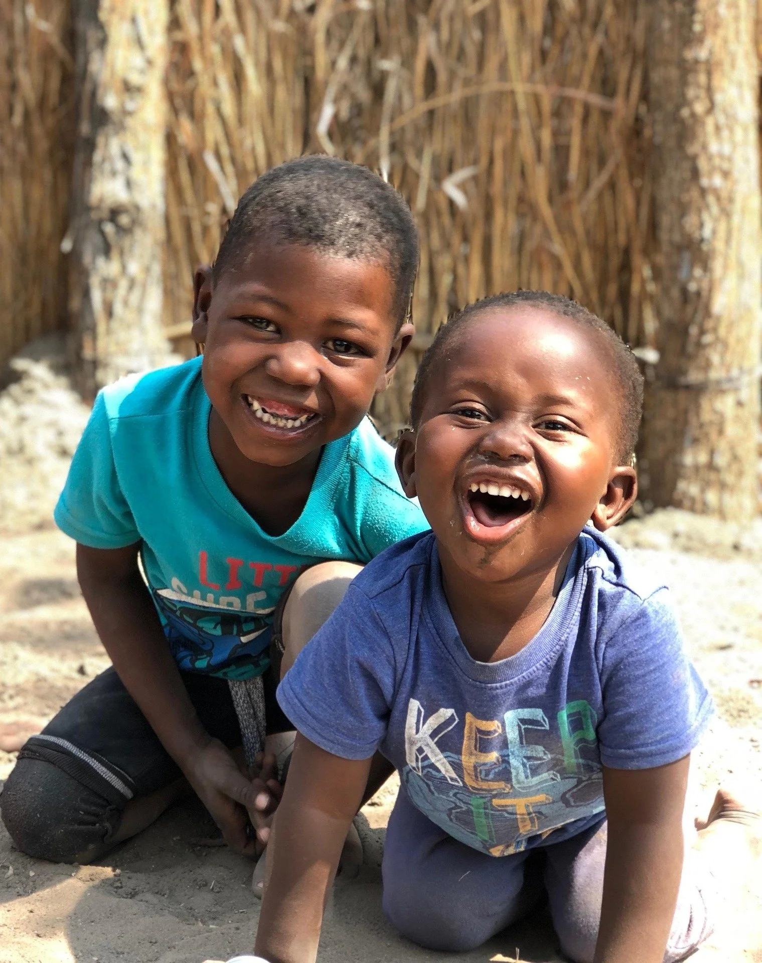 Two young boys smiling and playing on the sandy ground in front of a bamboo fence.