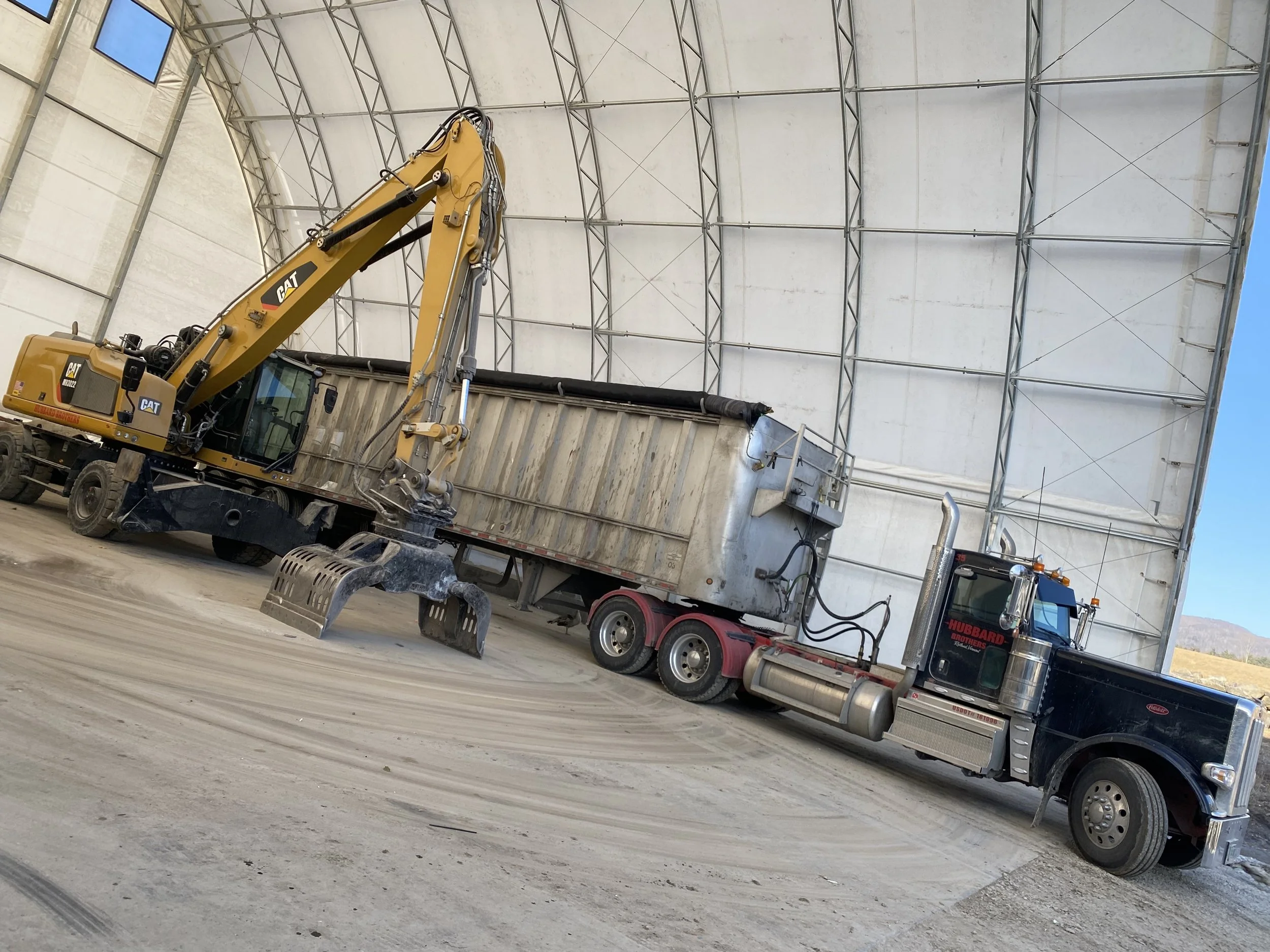 This image displays a material handler in the waste pit parked next to a hauling truck and trailer. The pit has been cleaned for the day and the final hauler loaded.