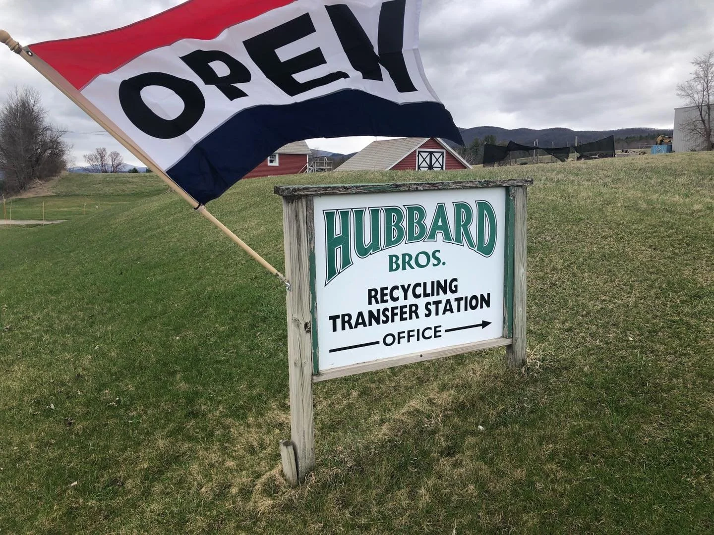 A sign for Hubbard Bros Recycling Transfer Station Office with a flag reading 'OPEX' attached above on a grassy area, with a cloudy sky and farm buildings in the background.