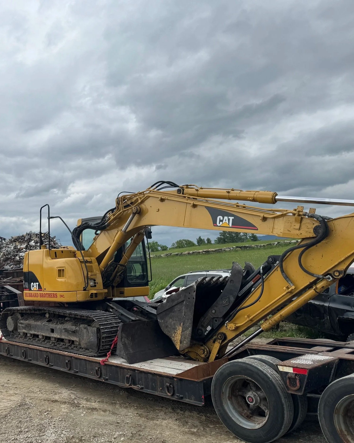 A yellow Caterpillar excavator loaded on a flatbed trailer on a gravel surface, with dark cloudy skies above and a grassy hill in the background.