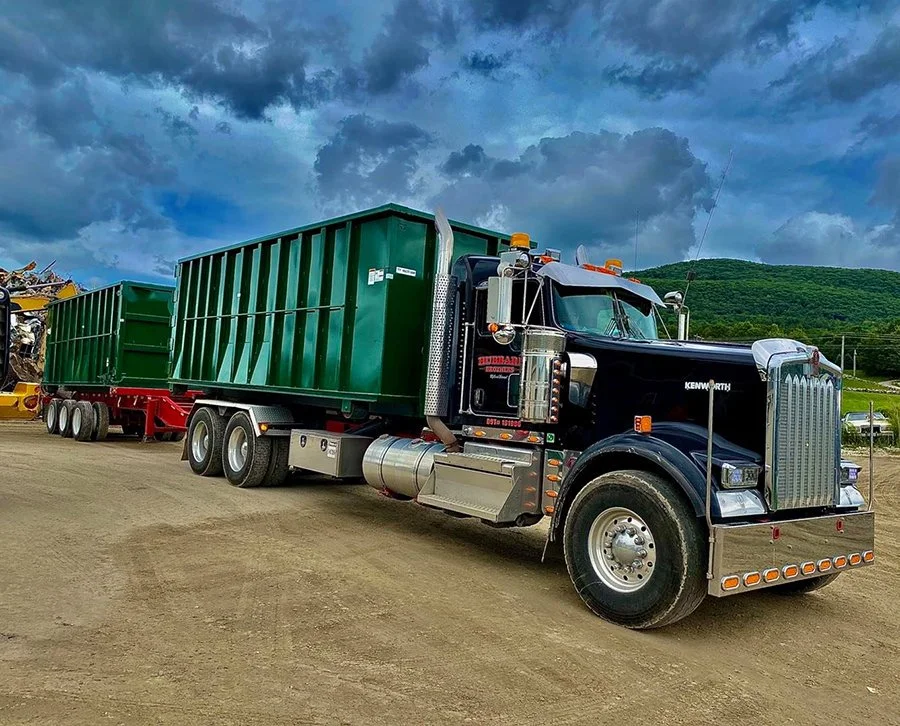 A black semi-truck with green containers attached, parked on a dirt lot under a cloudy sky, with green hills in the background.
