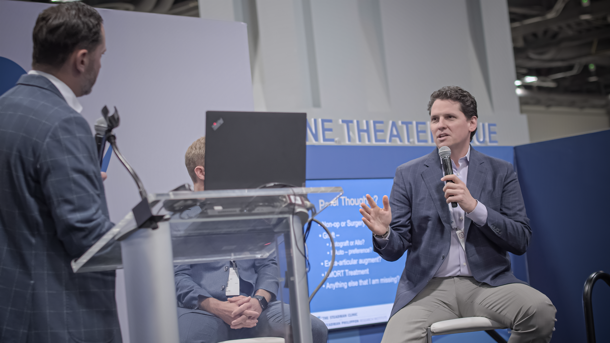 A man in a blue blazer and beige pants sitting on a chair holding a microphone, speaking to an audience. Two other men are standing nearby, one with a laptop, in front of a blue sign with white text.