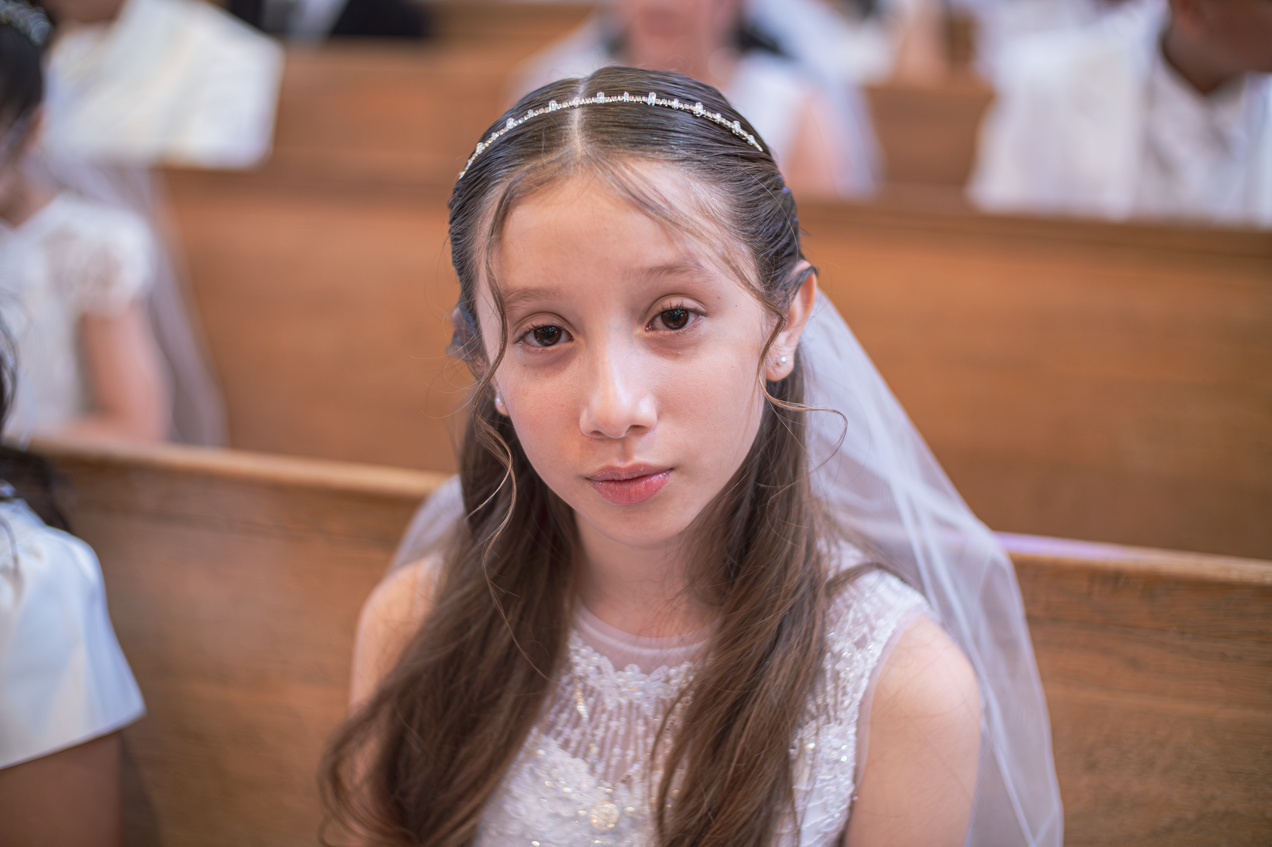 Young girl in a white dress and veil sitting in a church pew, likely at a wedding, looking directly at the camera.