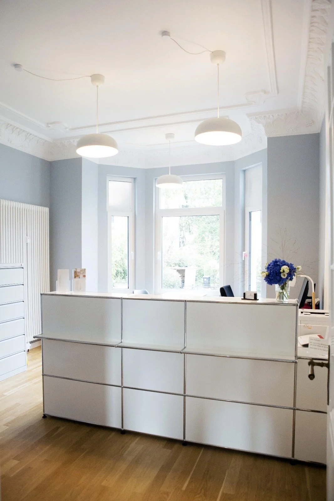 Ein heller Empfangstresen in einem Raum mit großen Fenstern, blauer Wandfarbe, weißen Deckenleuchten und einem blauen Blumenstrauß auf dem Empfangstresen.