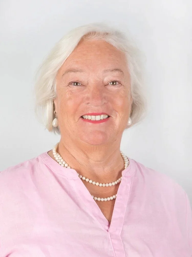 Portrait einer älteren Frau mit weißen Haaren, pinkem Hemd, Perlenkette und Ohrringen vor einem weißen Hintergrund.