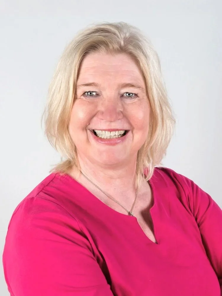 Lächelnde Frau mit blonden Haaren, trägt ein pinkes Oberteil und ein Halsketten, vor hellem Hintergrund.