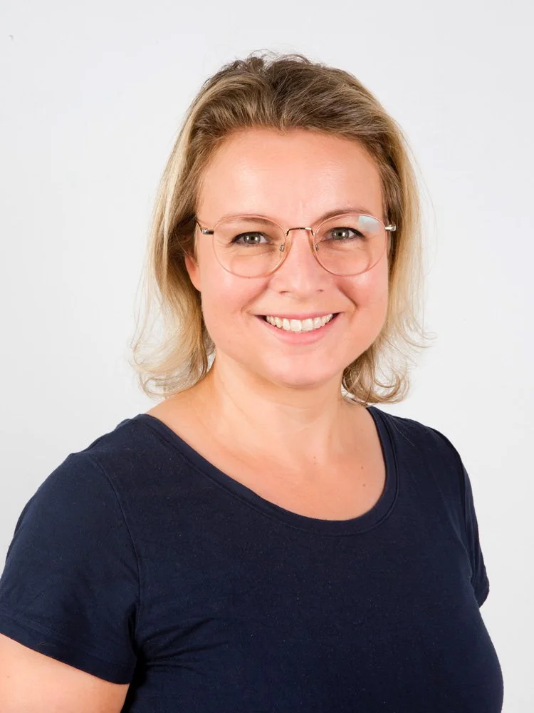 Eine lächelnde Frau mit Brille, kurze blonde Haare und dunkelblauem T-Shirt vor weißem Hintergrund.