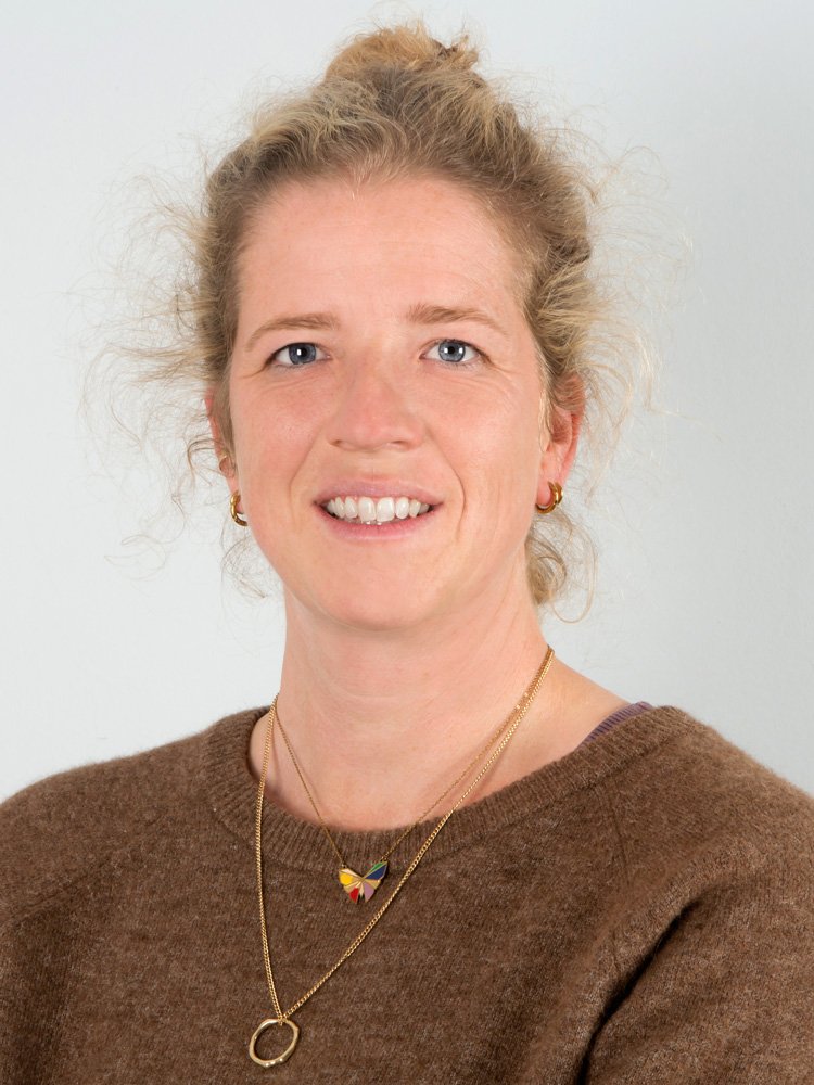 Porträt einer Frau mit braunem Pullover, goldenen Halsketten und Ohrringen, blonde lockige Haare, lächelnd, gegen weißen Hintergrund.