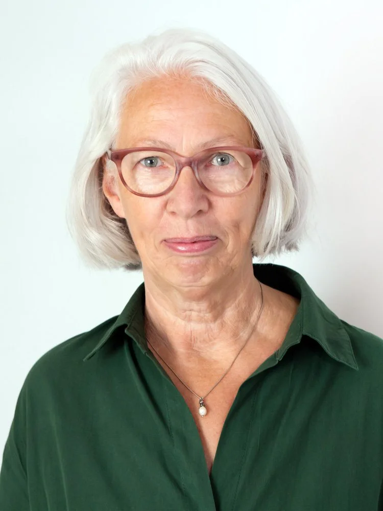 Porträt einer älteren Frau mit weißen Haaren und Brille, trägt ein grünes Hemd und eine Perlenkette, steht vor weißem Hintergrund.