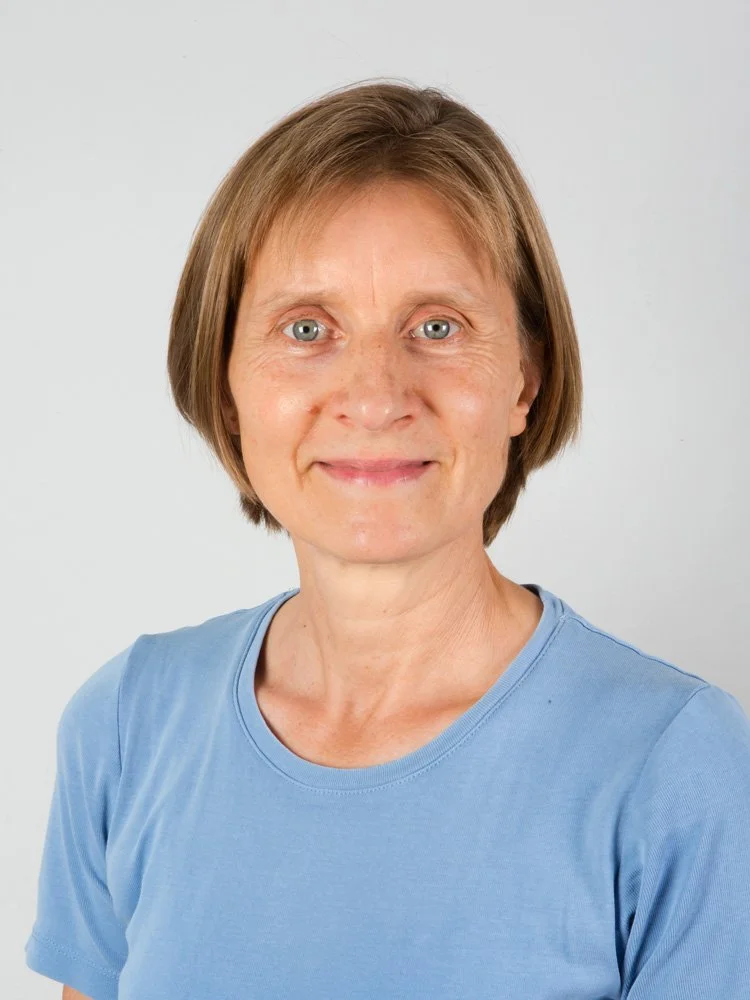Das Bild zeigt eine lächelnde Frau mit kurzen braunen Haaren, die ein hellblaues T-Shirt trägt, vor einem neutralen weißen Hintergrund.