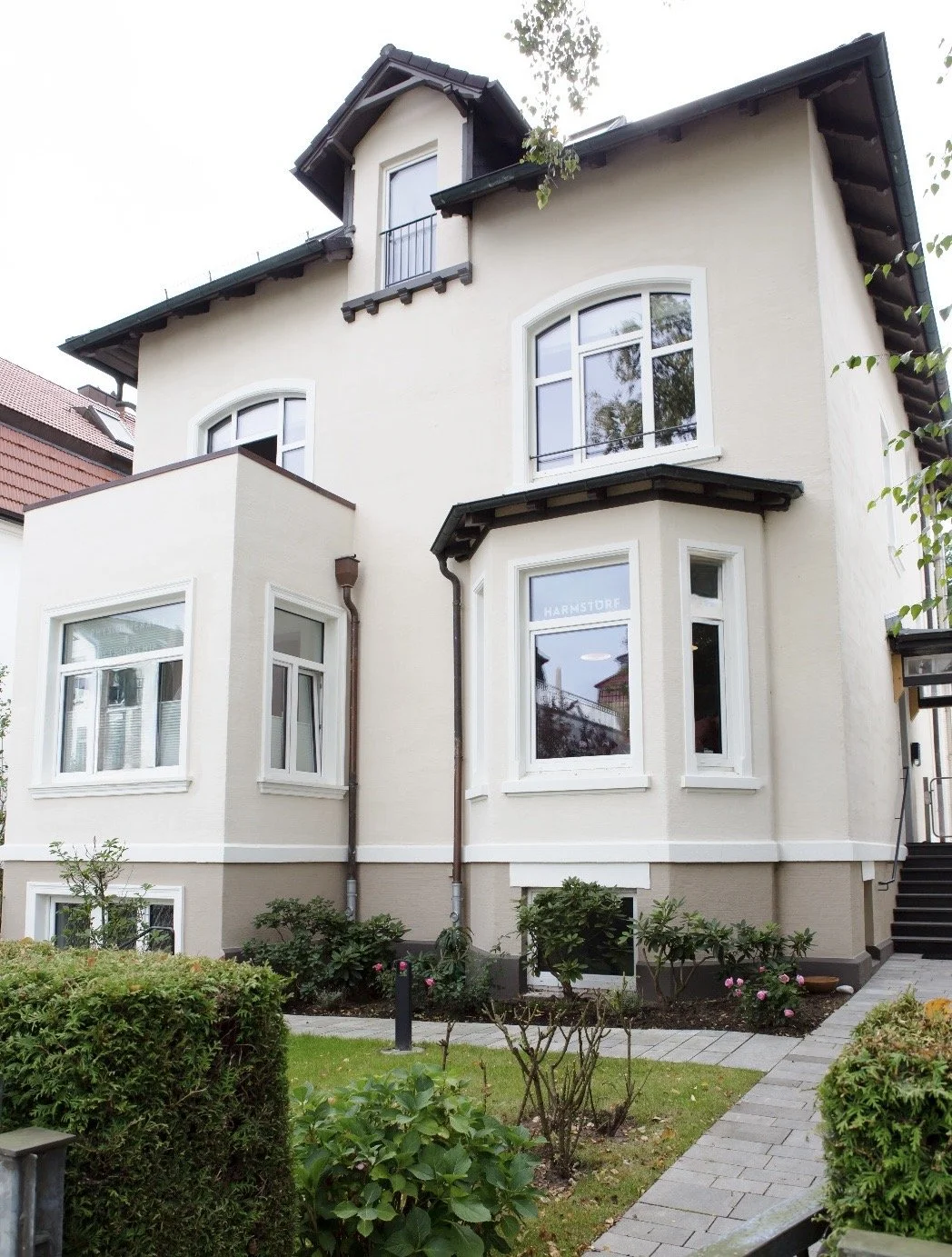 Mehrstöckiges Haus mit weißen Wänden, mehreren Fenstern und einem kleinen Garten mit Wegen, Pflanzen und Büschen.