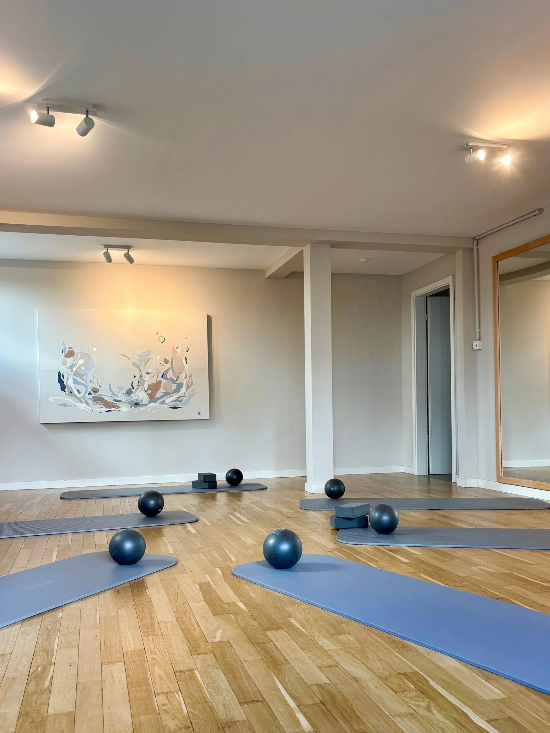 Fitnessraum mit Yoga-Matten, Gymnastikbällen und kleinen Gewichten auf Holzparkett, Wandbild an der Wand, Spiegel an der Seite, Deckenbeleuchtung.