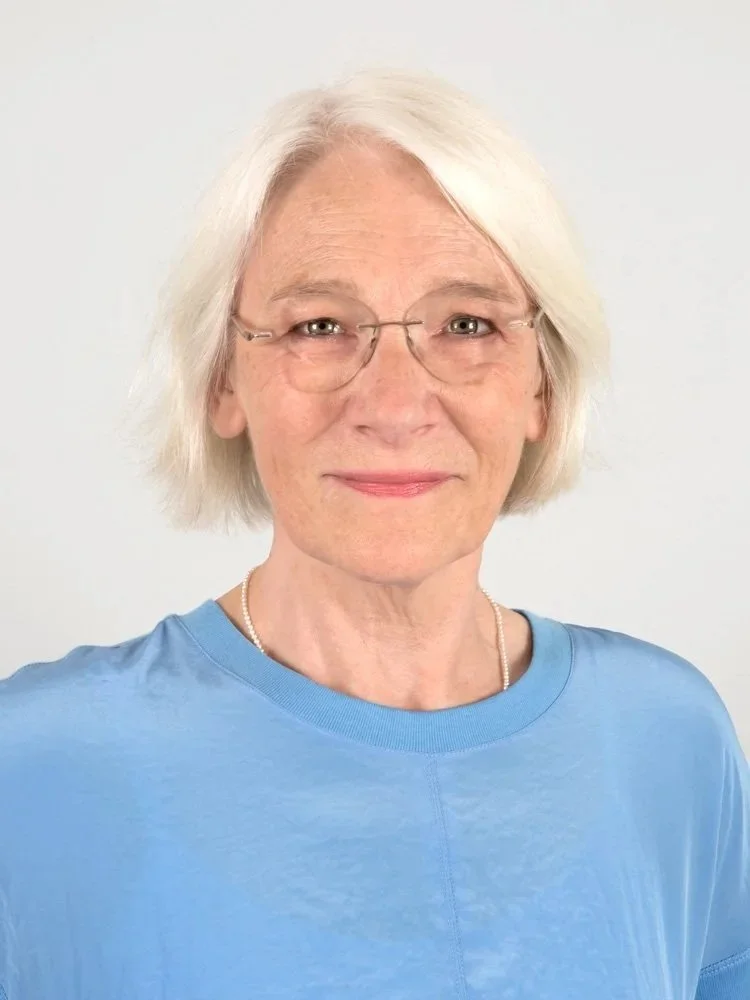 Nahaufnahme einer älteren Frau mit weißen Haaren und Brille, die ein blaues T-Shirt trägt, vor hellem Hintergrund.