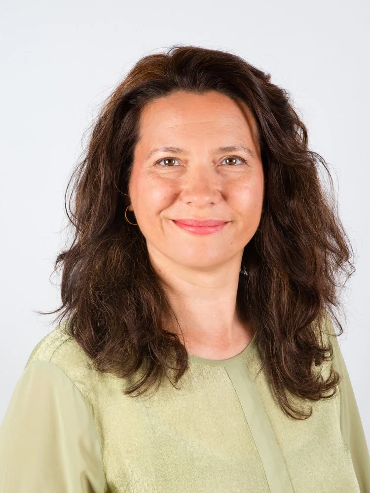 Porträt einer lachenden Frau mit braunen, lockigen Haaren und dezentem Make-up, trägt eine grüne Bluse vor einem weißen Hintergrund.