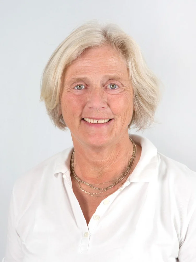 Ein älterer Frau mit kurzen, blondgrauen Haaren, die lächelnd in die Kamera schaut, trägt eine weiße Bluse und mehrere goldene Halsketten.