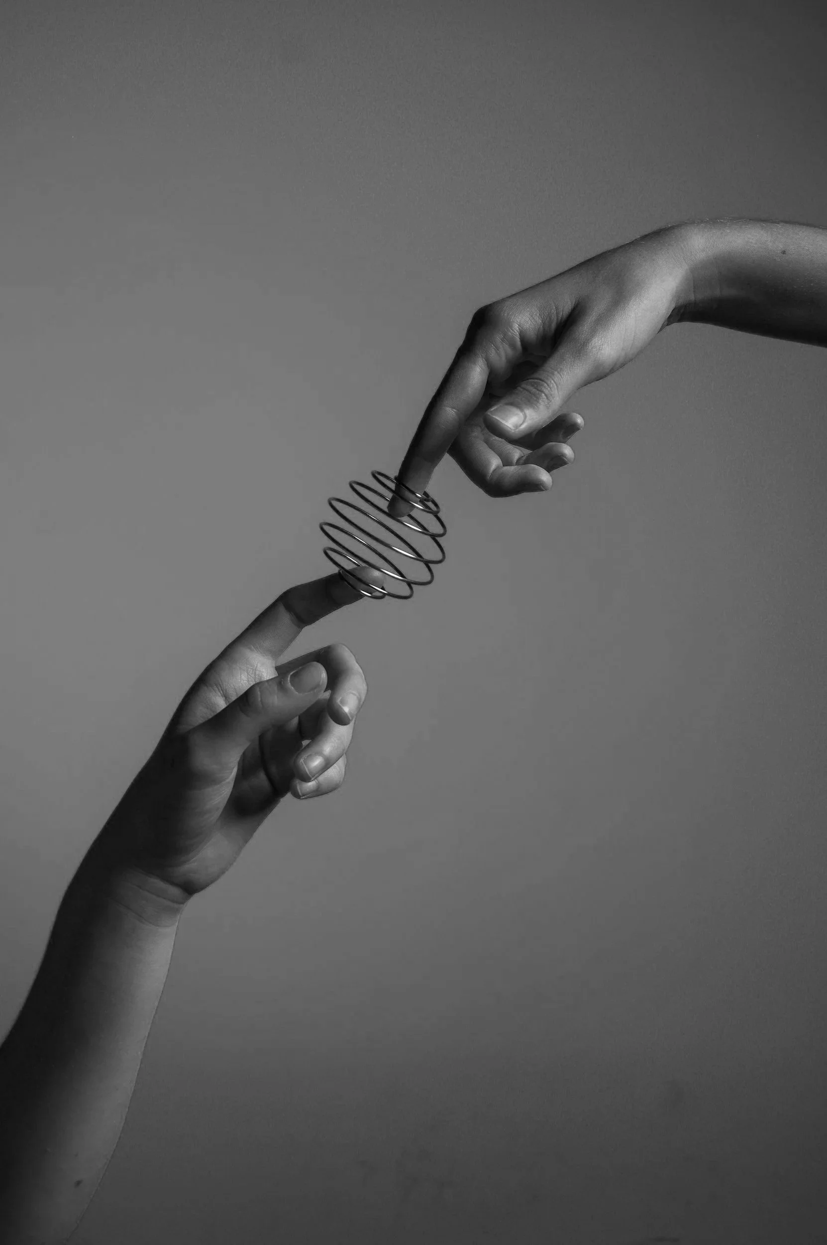 Two hands, one above the other, touching with a spring between their index fingers, in a black-and-white photo.