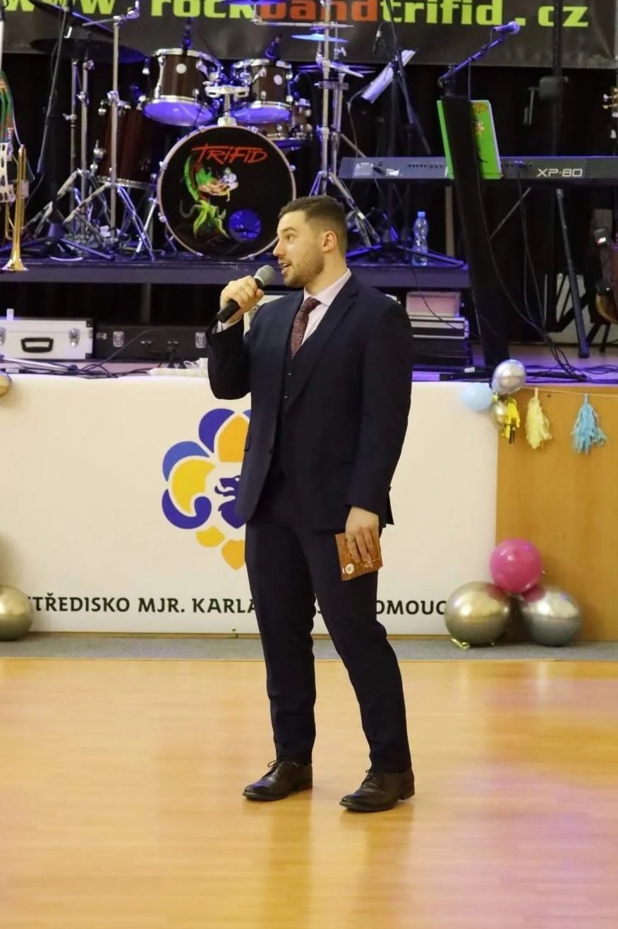Mladý muž, který je moderátorem. Má na sobě tmavě modrý navy oblek. V ruce drží mikrofon a vypráví o budoucnosti.