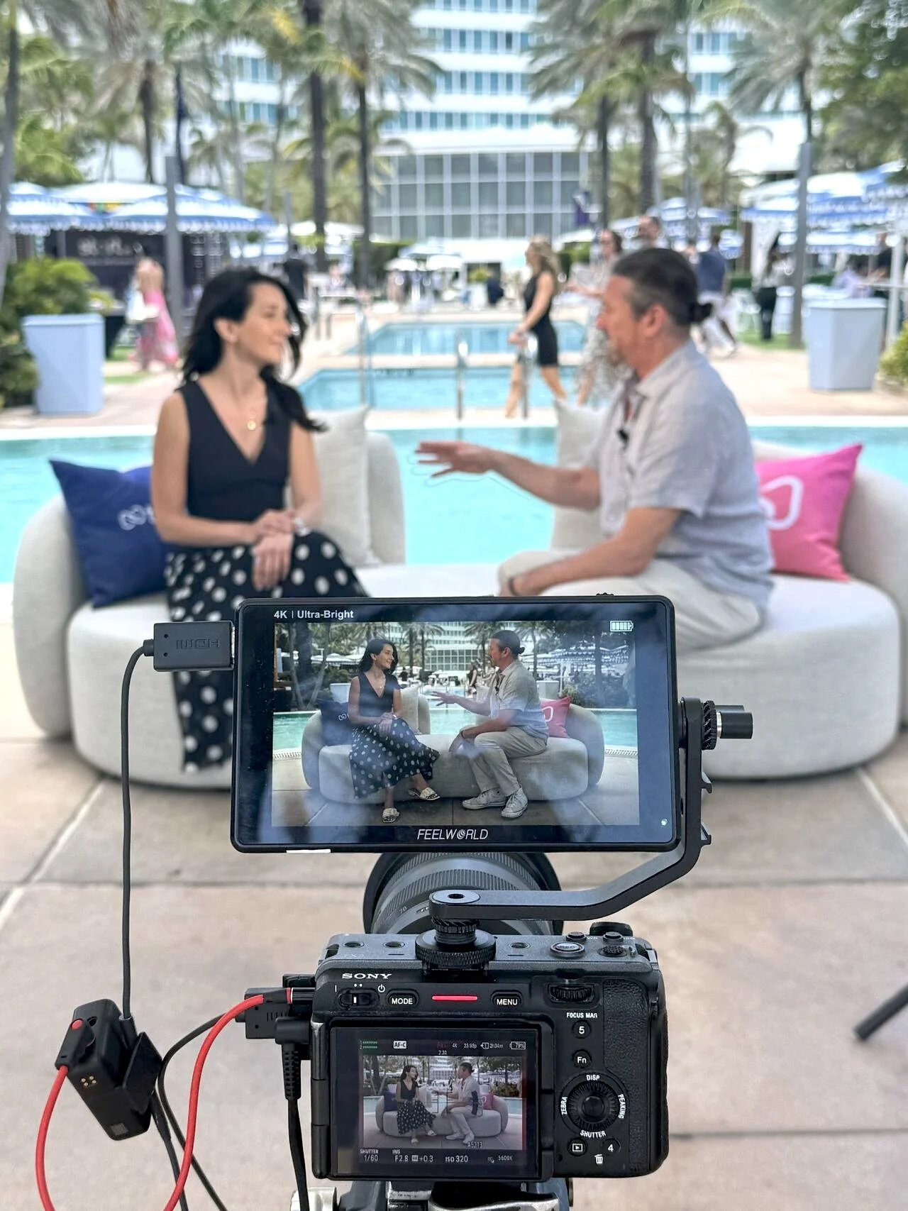 Katie Secret and Brian Monahan sit on a white sofa in front of a pool during an interview at Possible Miami 2025; the scene is being filmed with a professional camera showing the two people on its screen.