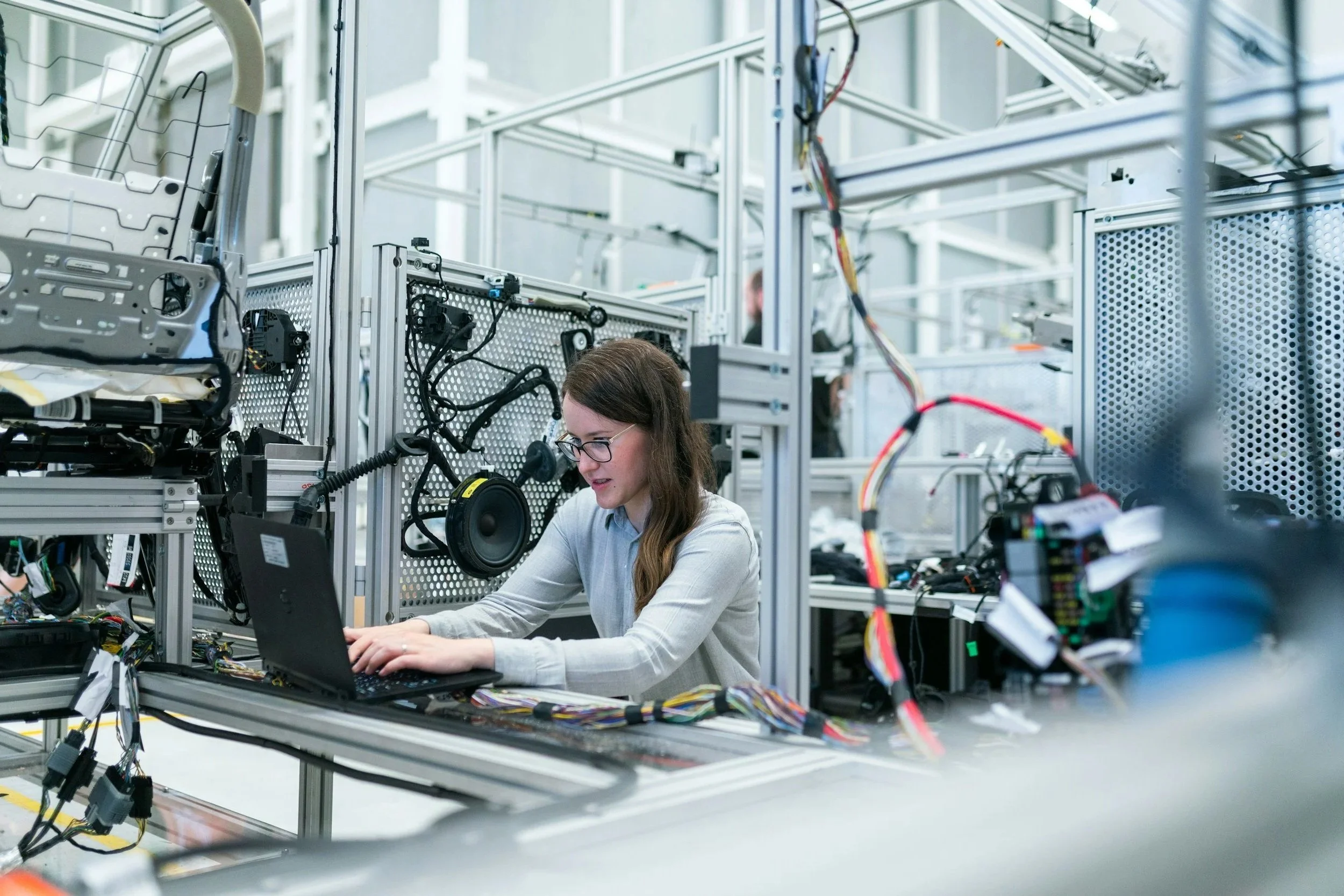 Mujer trabajando en una mesa con componentes electrónicos y una laptop en un entorno de laboratorio o taller tecnológico.