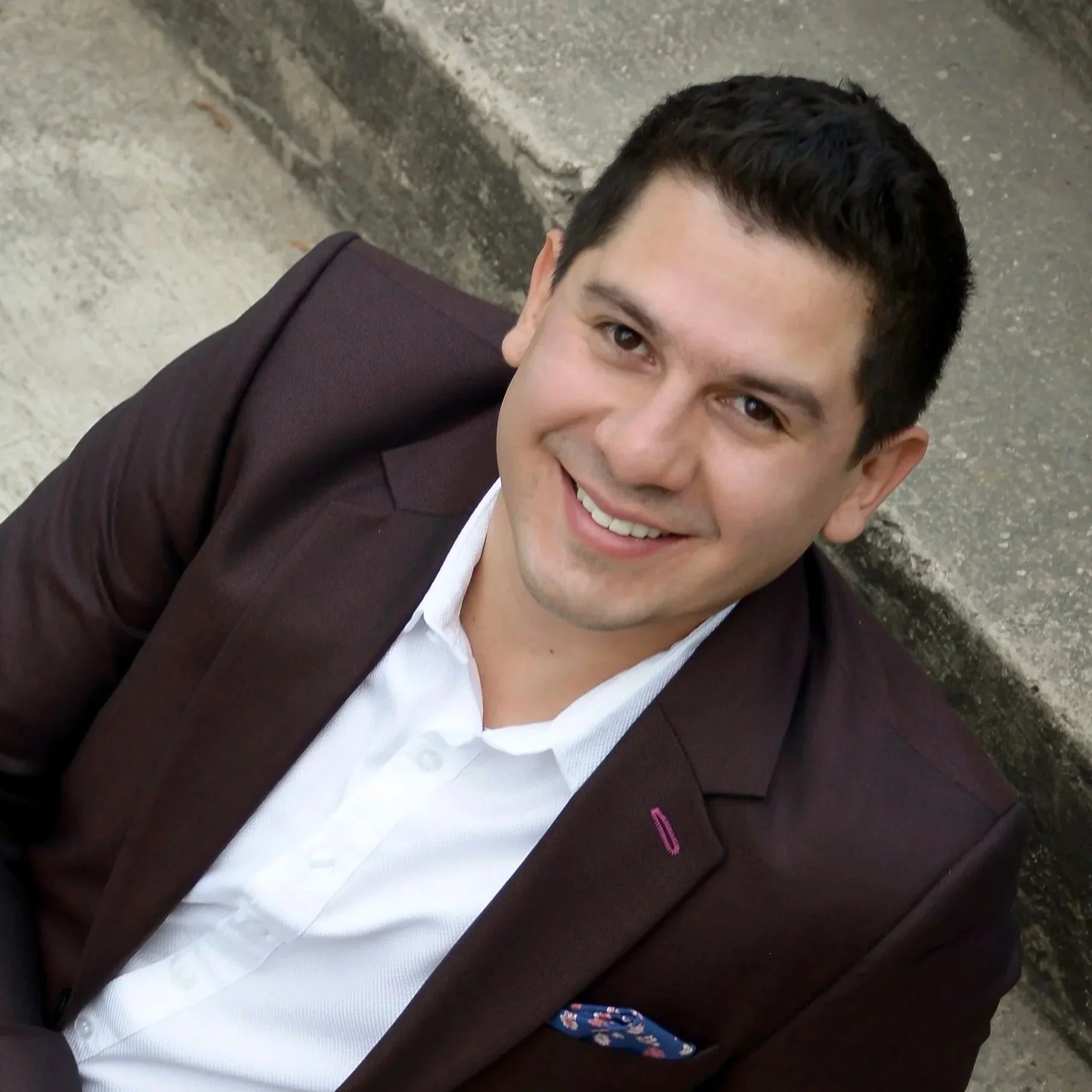 Joven sonriendo, usando chaqueta marrón y camisa blanca, sentado en la calle.
