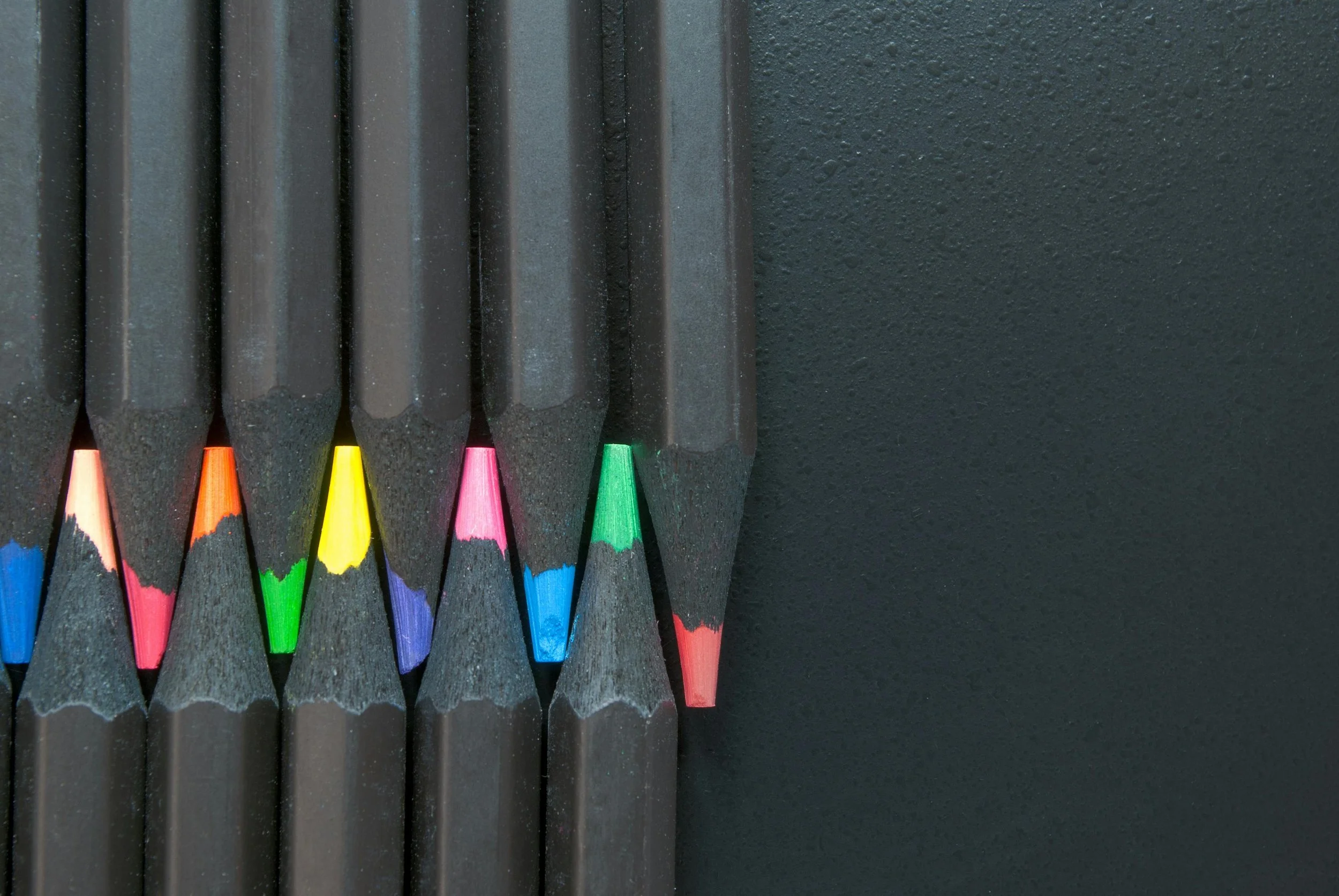 A row of sharpened colored pencils with black bodies, arranged against a textured black background.