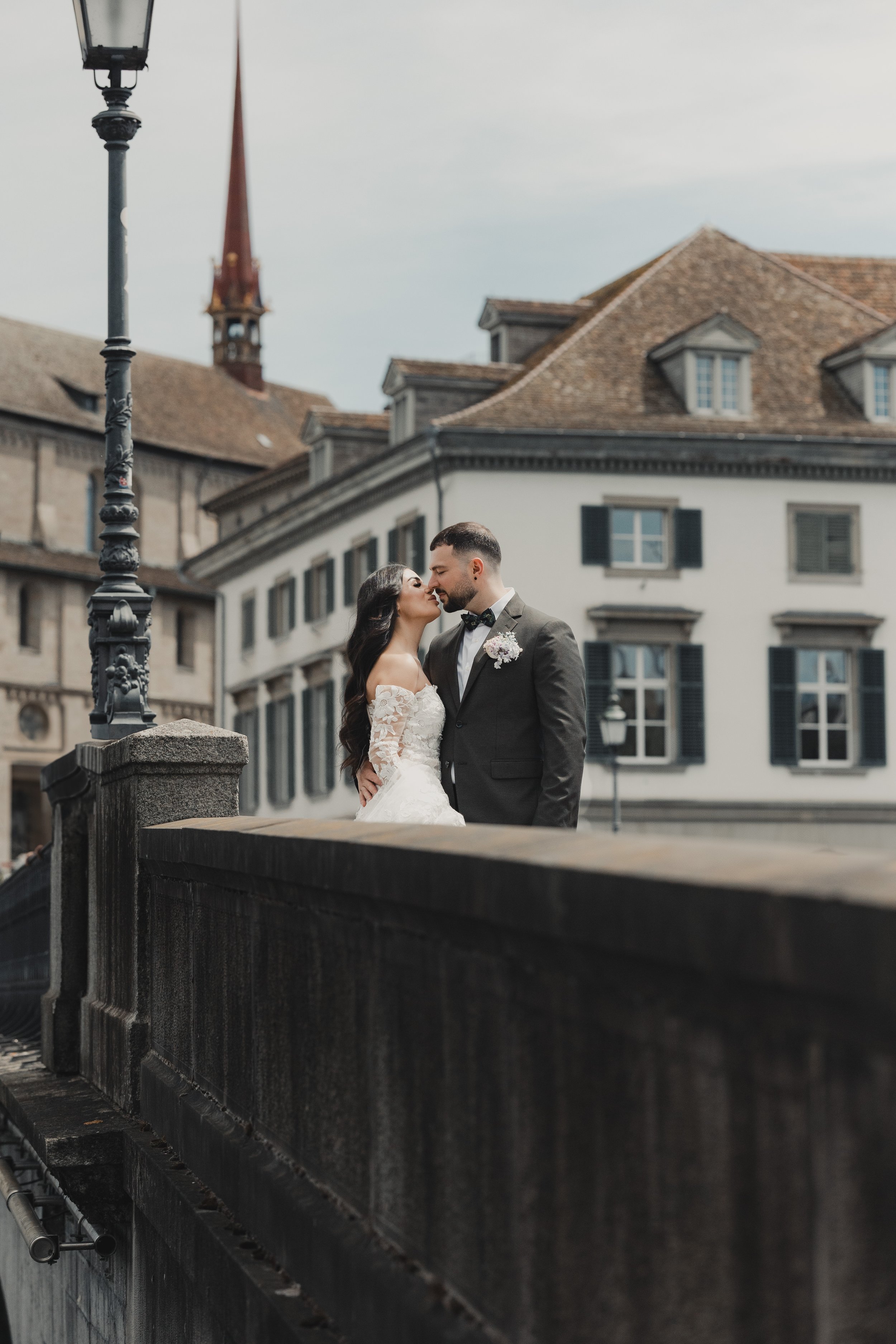 Ein Brautpaar küsst sich auf einer Brücke in einer Stadt mit historischen Gebäuden und einem Kirchturm im Hintergrund.