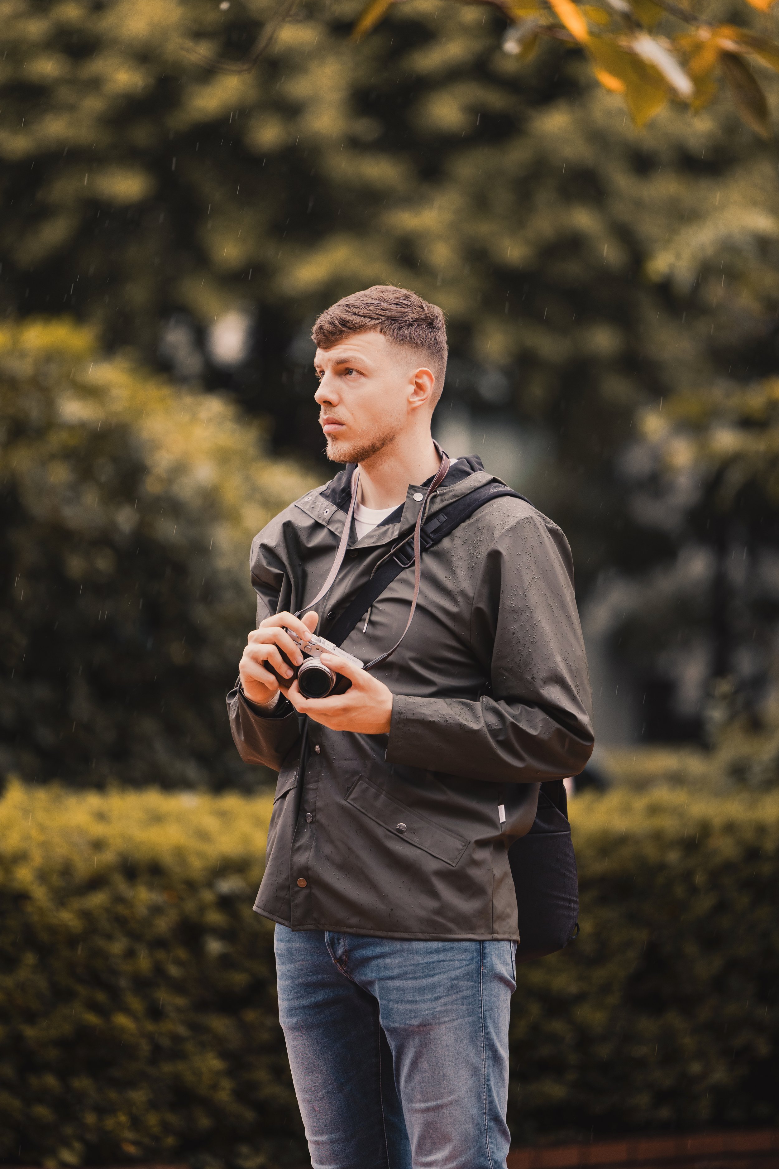 Junger Mann mit Kamera in der Hand, trägt eine Jacke und einen Rucksack, steht im Regen in einem Park mit bunter Laubbedeckung.