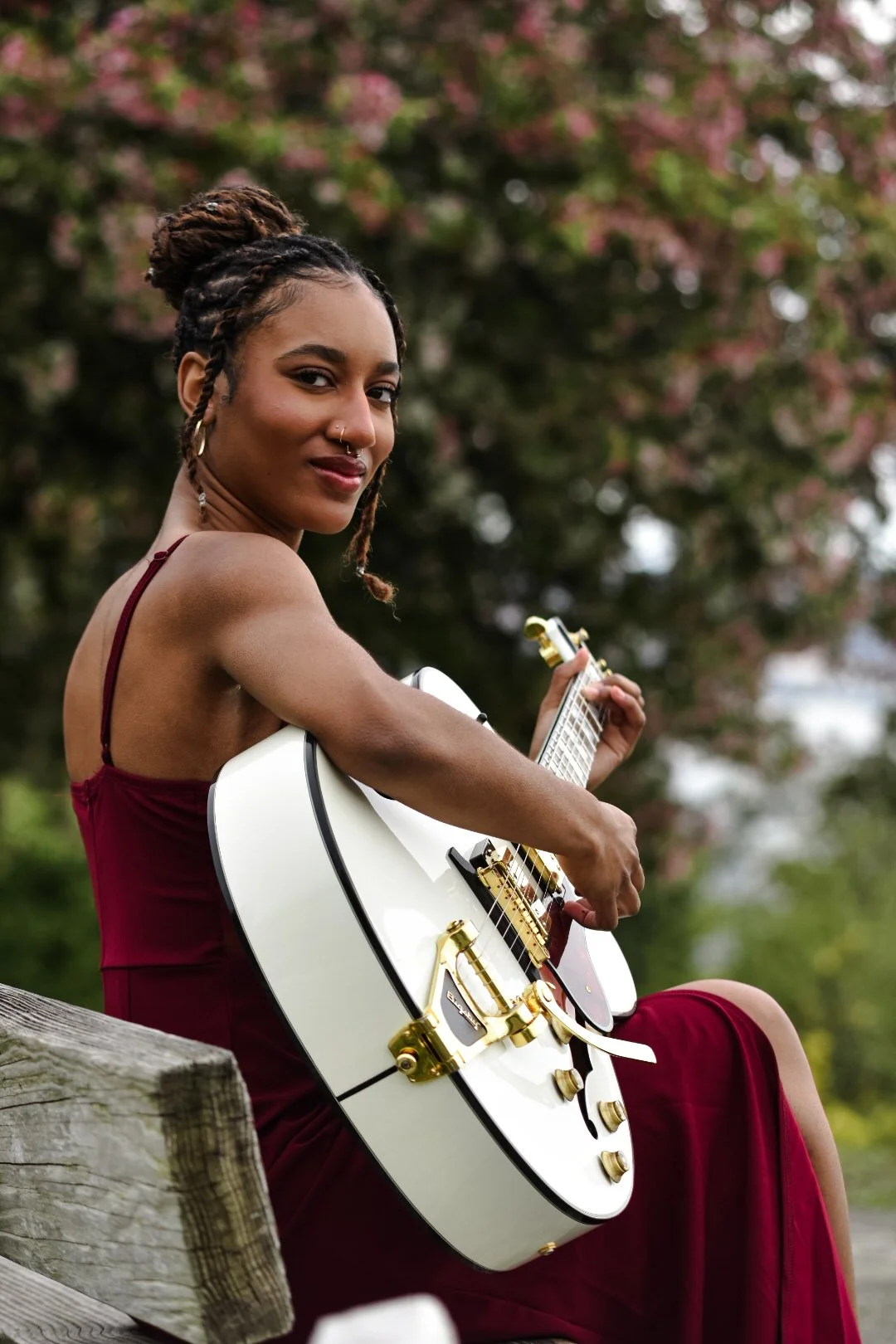 A woman with dreadlocks and a nose piercing sitting on a wooden bench outdoors, holding a white electric guitar with gold hardware, and looking at the camera with a slight smile.