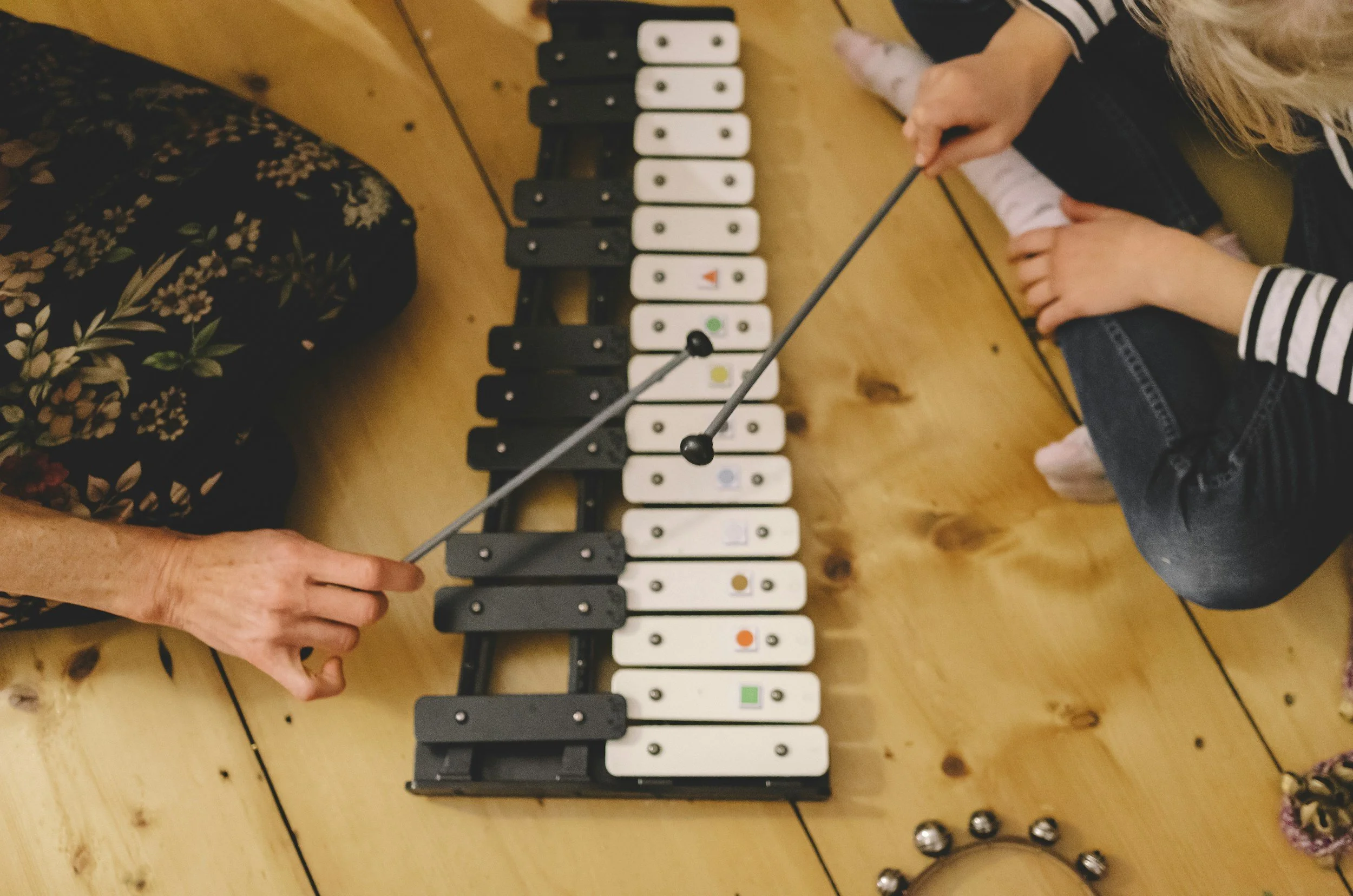 Two people playing a small xylophone with black and white keys and colorful dots, using mallets, on a wooden table. One person is sitting on the right side, and the other is reaching from the left side.