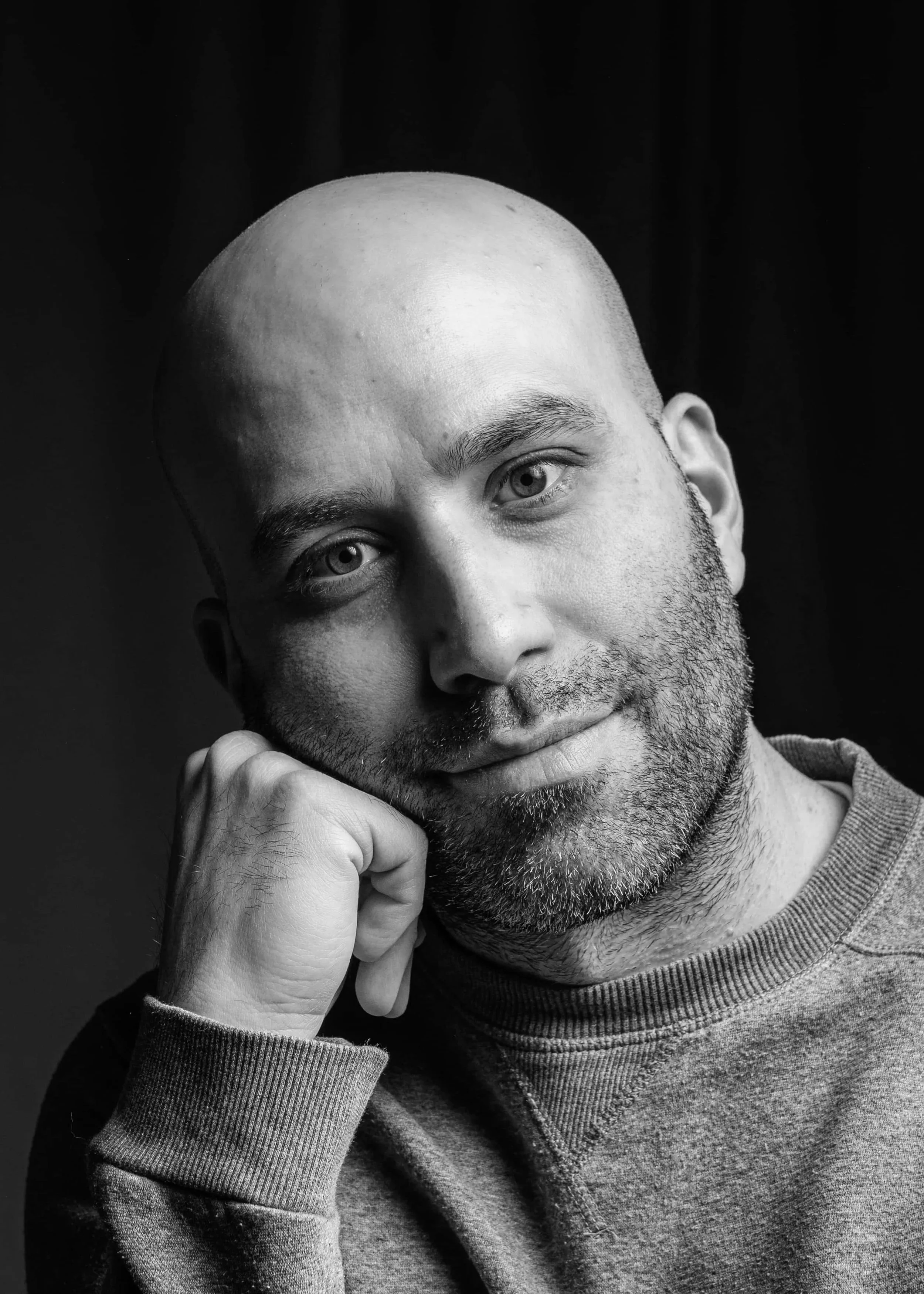 Black and white portrait of a man with a shaved head, beard, and expressive eyes, resting his chin on his right hand, wearing a sweatshirt.