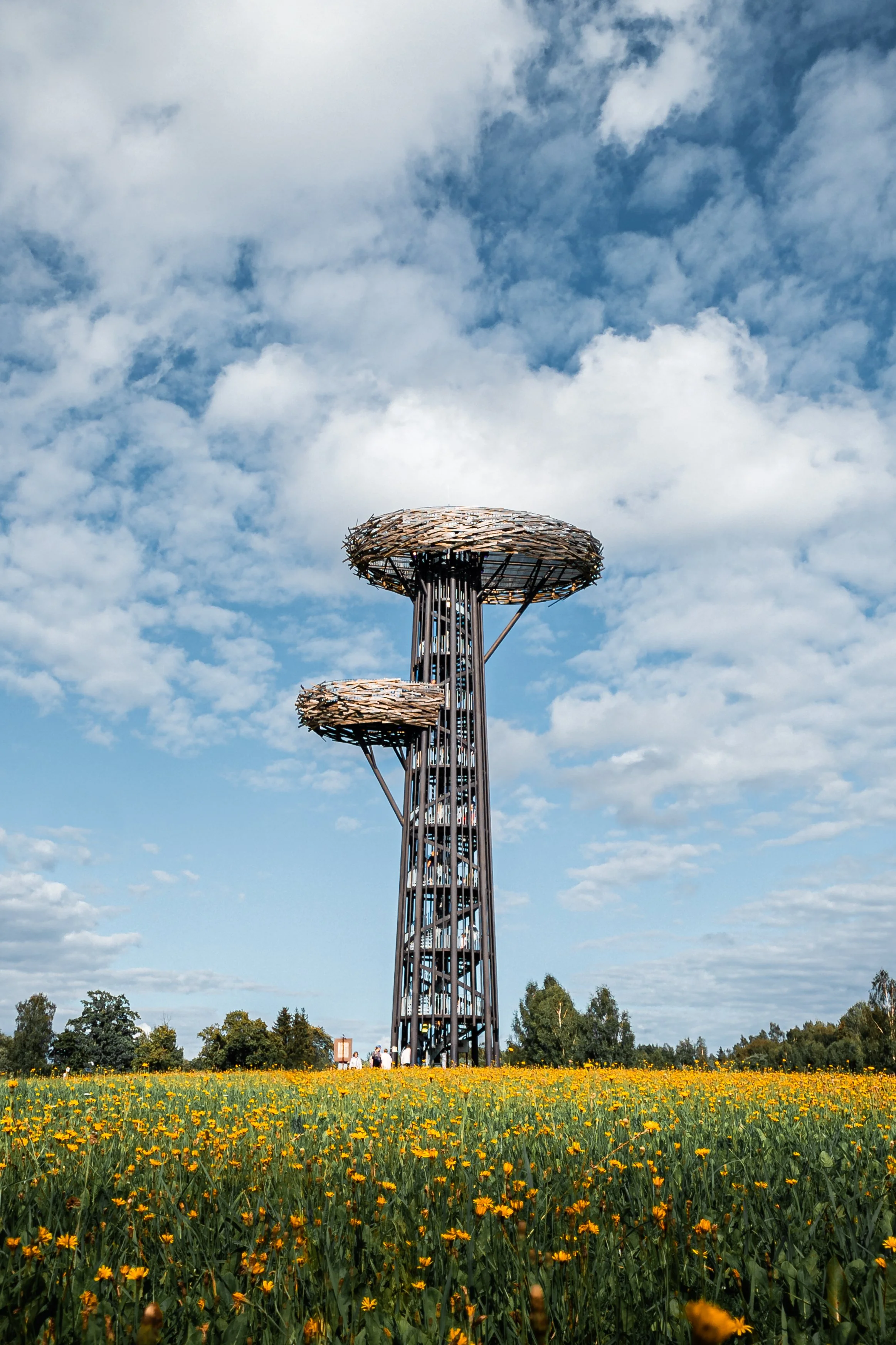 A tall tree-like structure with three circular platforms in a field of yellow flowers, under a partly cloudy sky.