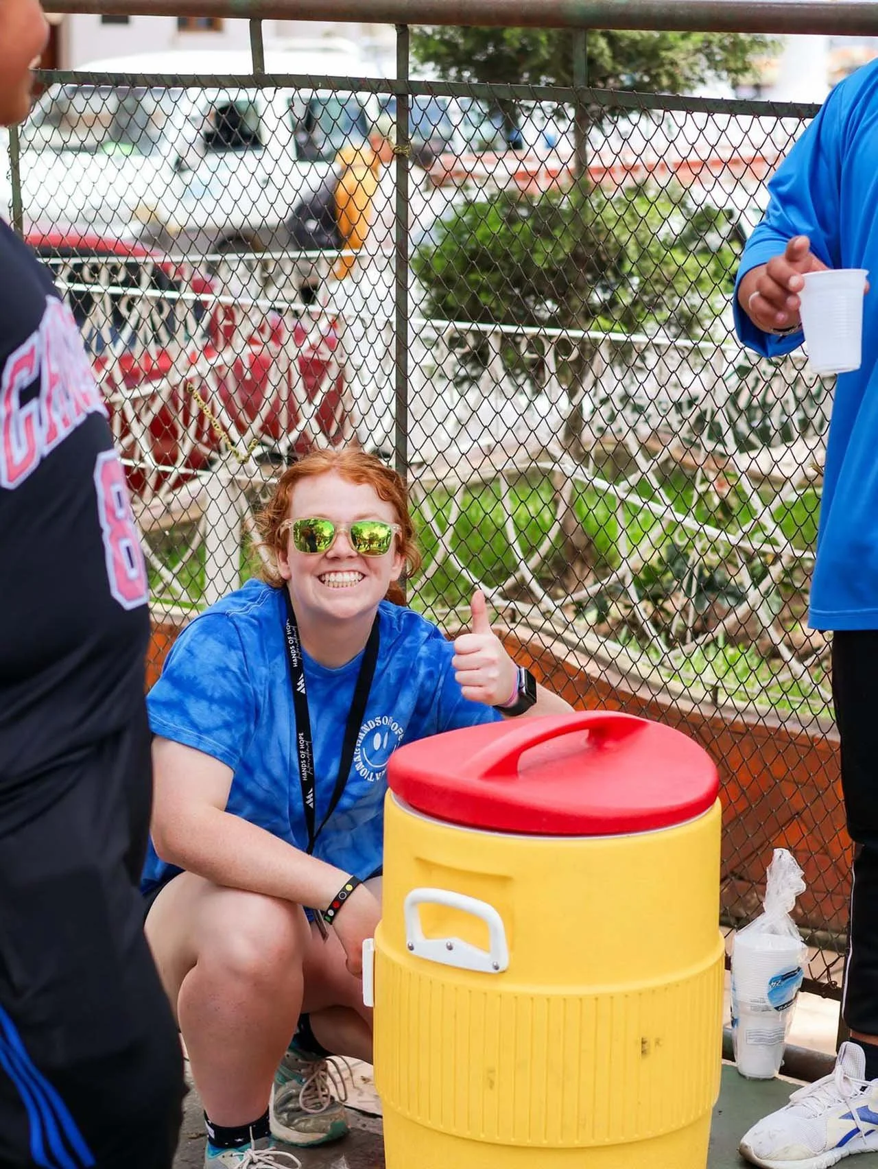 Young woman with red hair, sunglasses, and a blue T-shirt giving a thumbs-up while sitting near a yellow water cooler at an outdoor event.