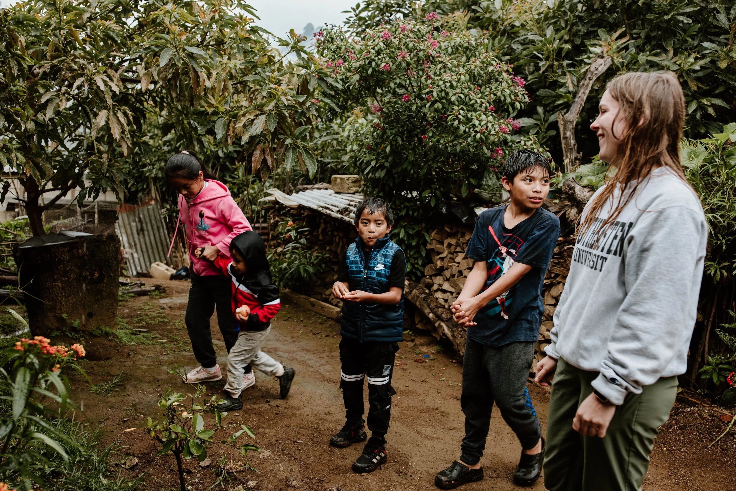 A group of five children and an adult woman walking outdoors on a dirt path surrounded by greenery and plants.