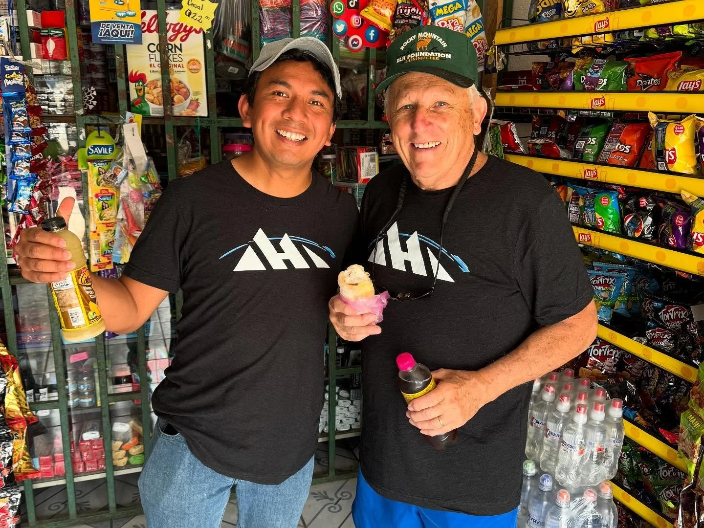 Two men smiling inside a convenience store, one holding a bottle of juice and the other holding an ice cream cone, surrounded by snack shelves and bottled water.