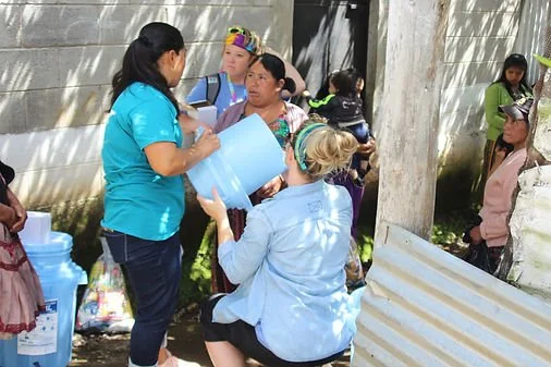 A group of women and children are gathered outdoors near a fence, with some sitting and others standing. The scene suggests a community or family event.