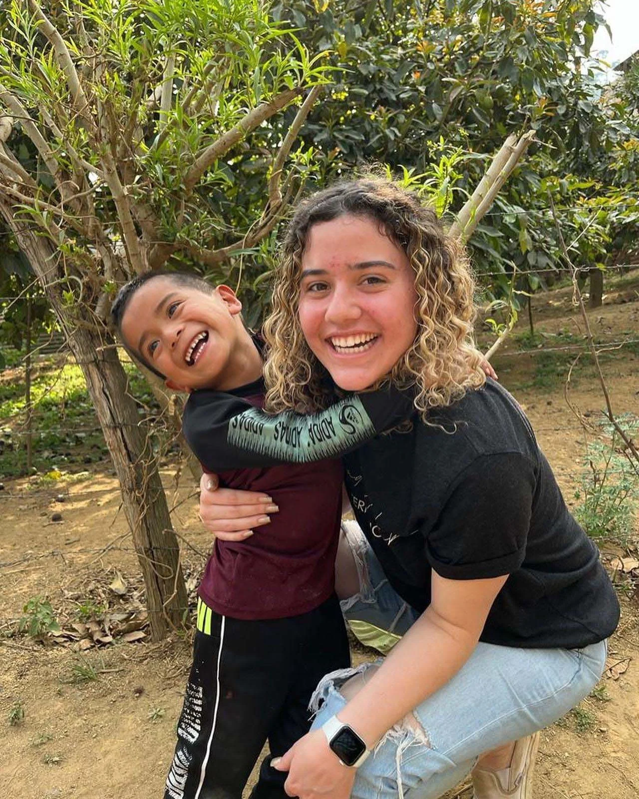 A woman crouching next to a young boy, both smiling and hugging outdoors with trees and plants in the background.