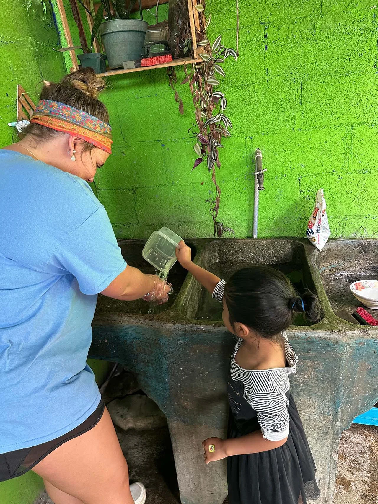A woman and a girl washing something in a large, concrete sink. The woman is wearing a colorful headband and a blue t-shirt. The girl has dark hair tied back and is wearing a black skirt and striped top.