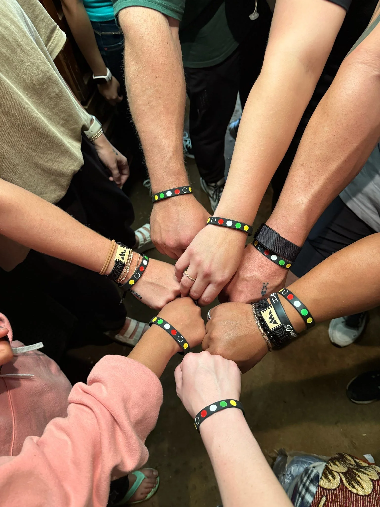 A group of diverse people with different skin tones and ages showing their wrists with matching black bracelets featuring colorful dots, forming a circle with their fists joined in the center.