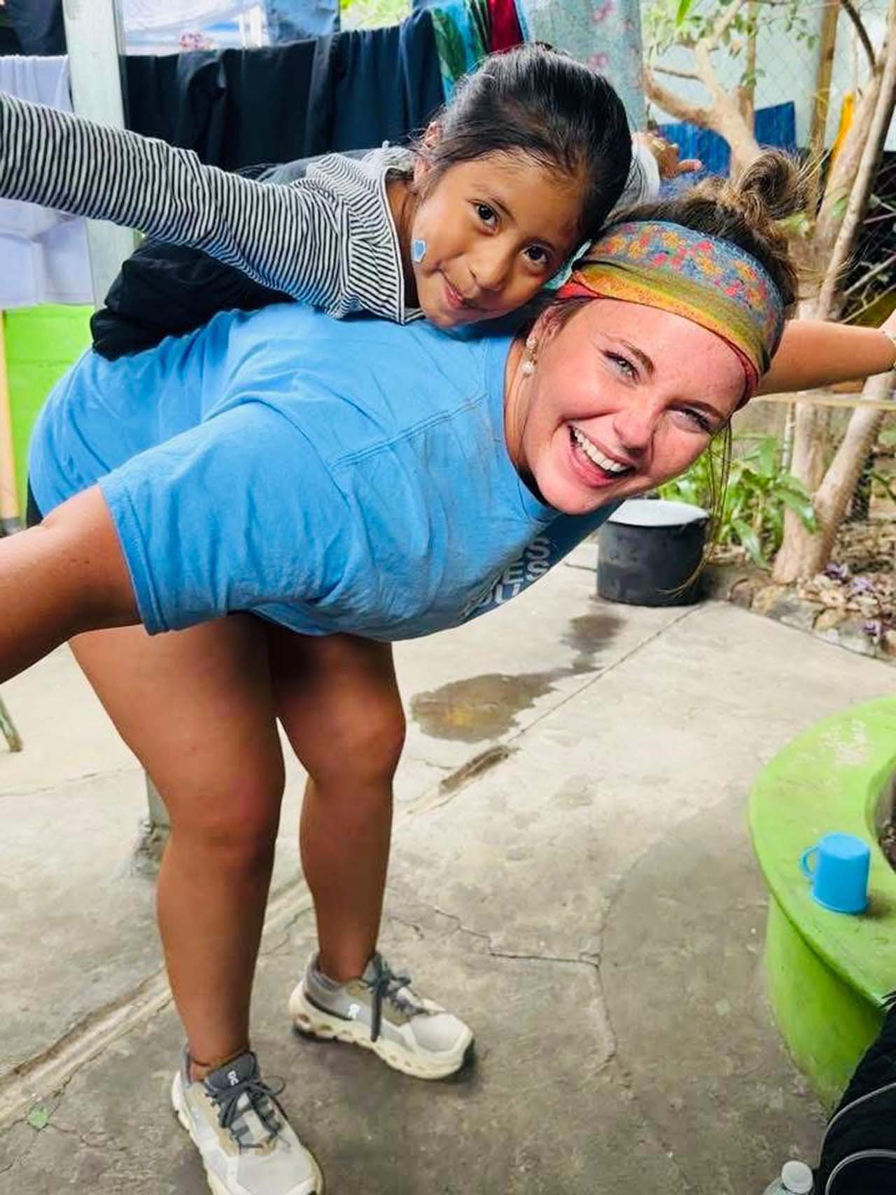 A woman and a young girl are playing together outdoors, with the woman bending forward and the girl lying on her back on the woman's back, both smiling.