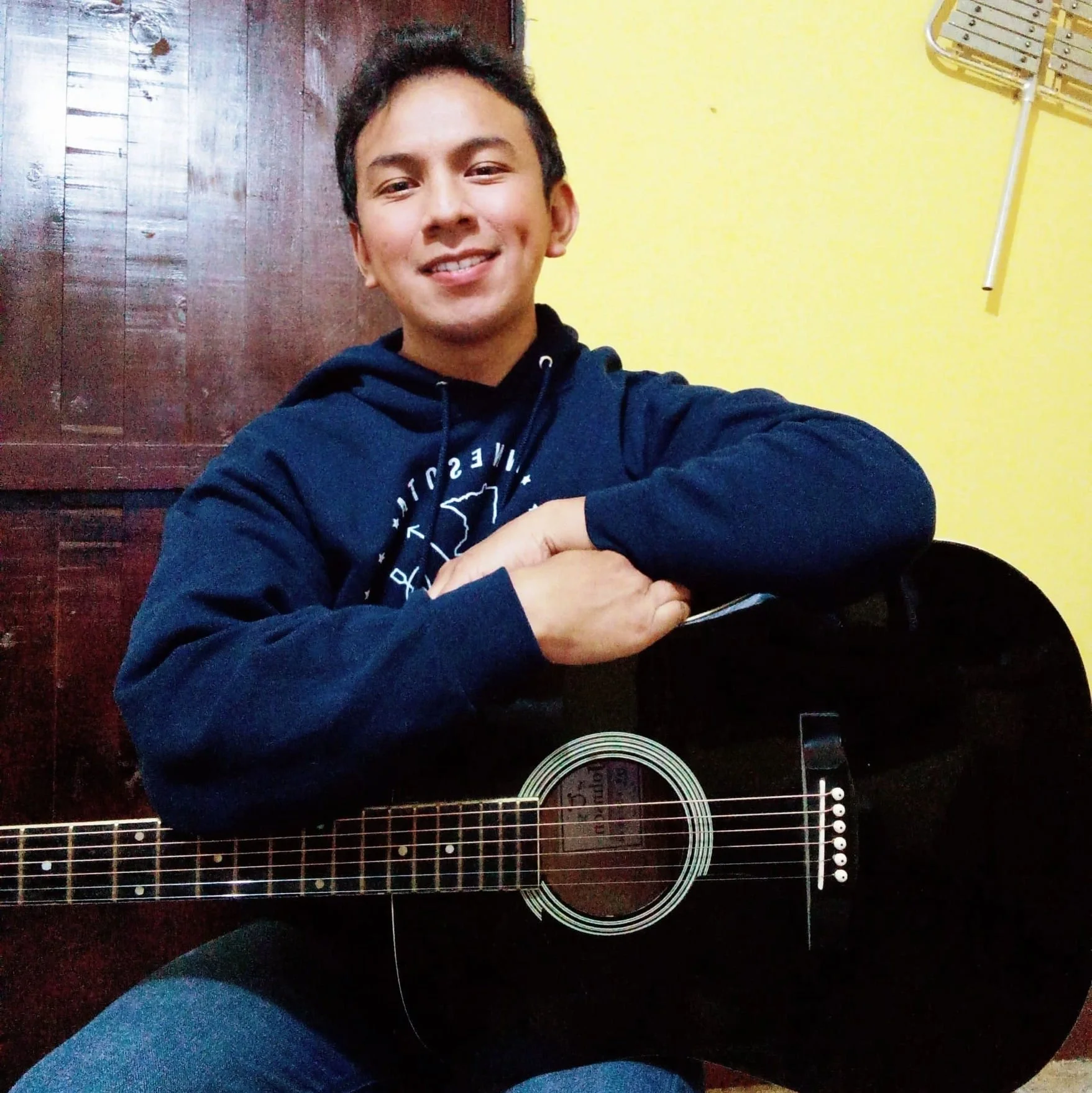 A young man smiling while sitting with an acoustic guitar, wearing a navy hoodie, in a room with a yellow wall and wooden door.