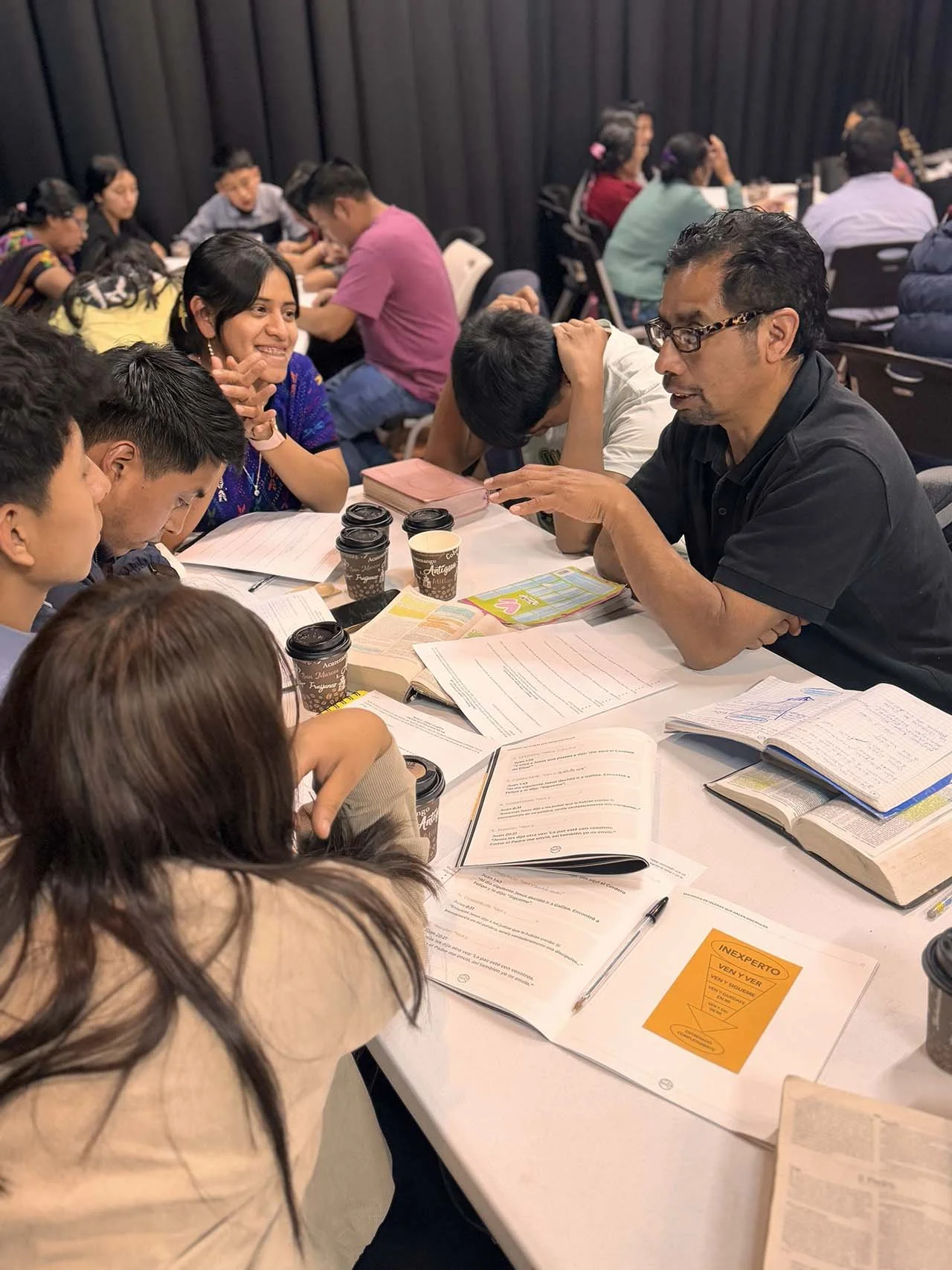 A group of diverse people engaged in a discussion around a round table, with books and papers open, cups of coffee, and a black curtain in the background.