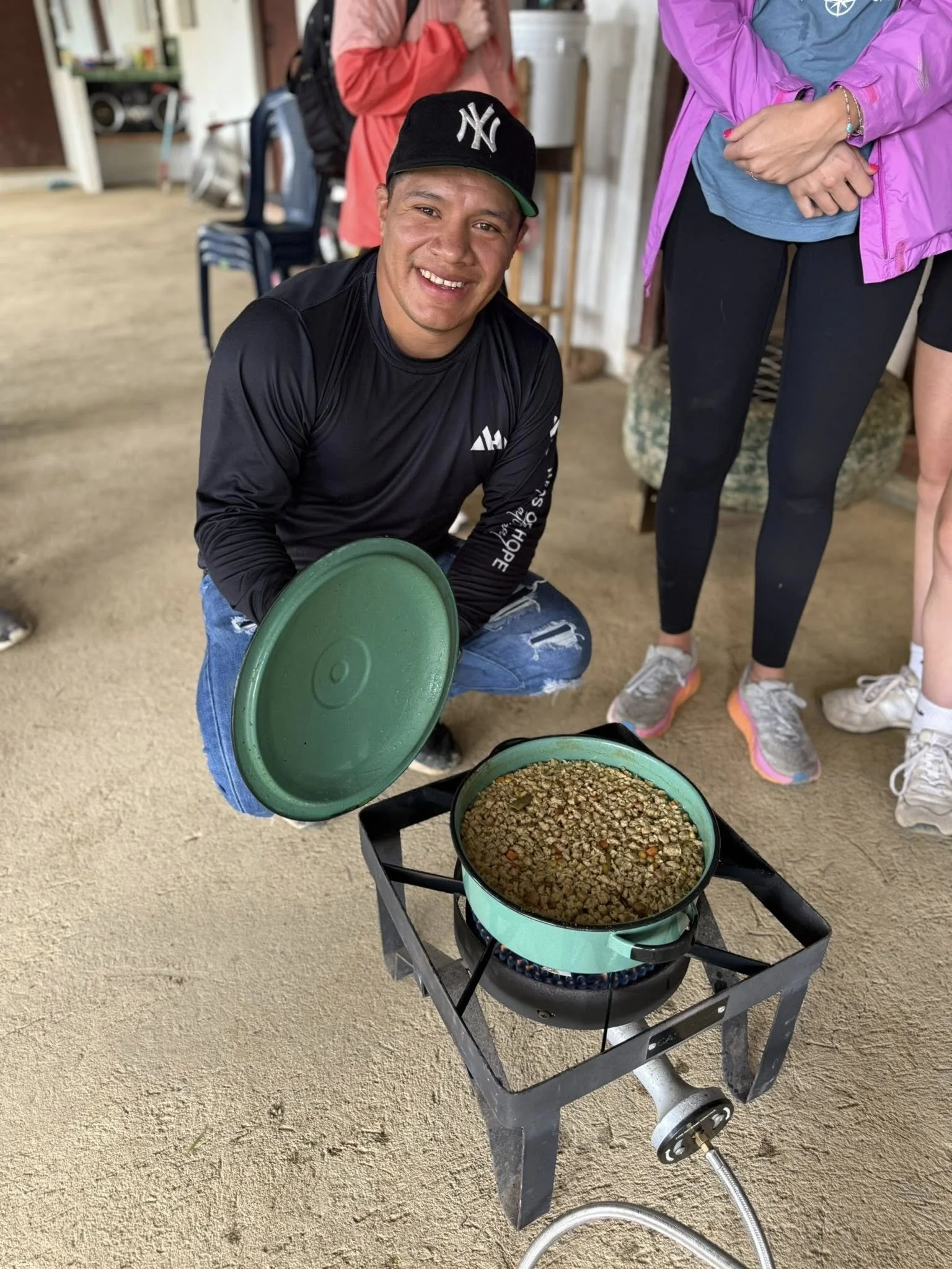 A smiling young man in a black long sleeve shirt and black New York Yankees cap squatting beside a portable propane stove with a pot of cooked lentils, holding a green lid in his right hand. Two women stand nearby, one in black leggings and pink jacket, another in black shorts and white sneakers.